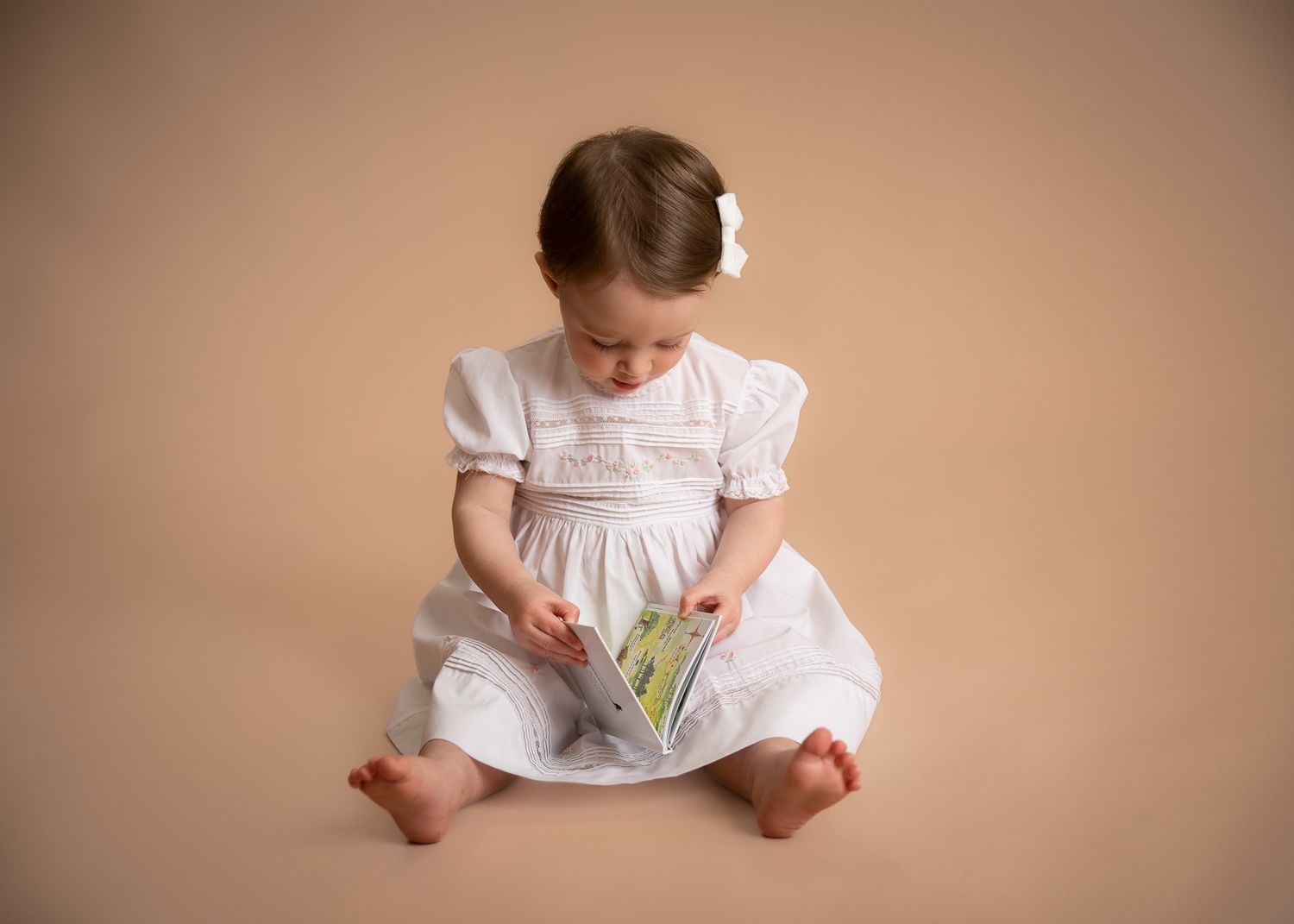Cute toddler girl in white smocked dress reading a book on beige studio background.