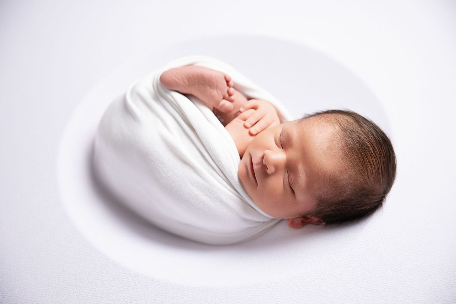 Peaceful newborn baby wrapped in white swaddle sleeping on white background in classic posed photo.