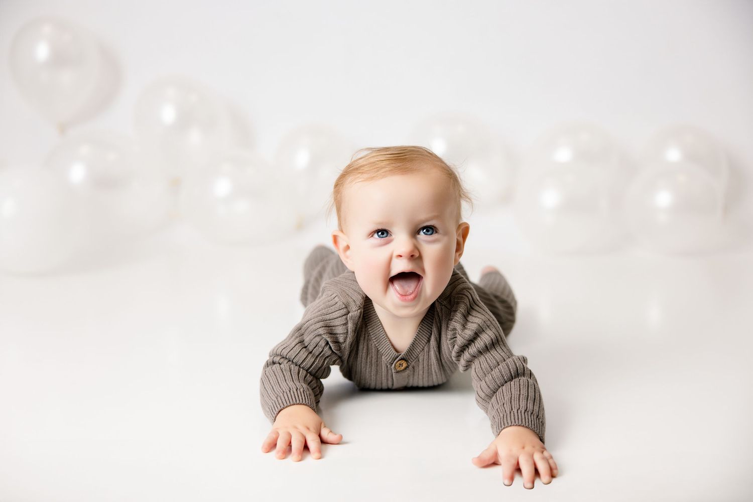 A happy baby in a brown knit outfit lies on a white surface, smiling with clear balloons in the background.