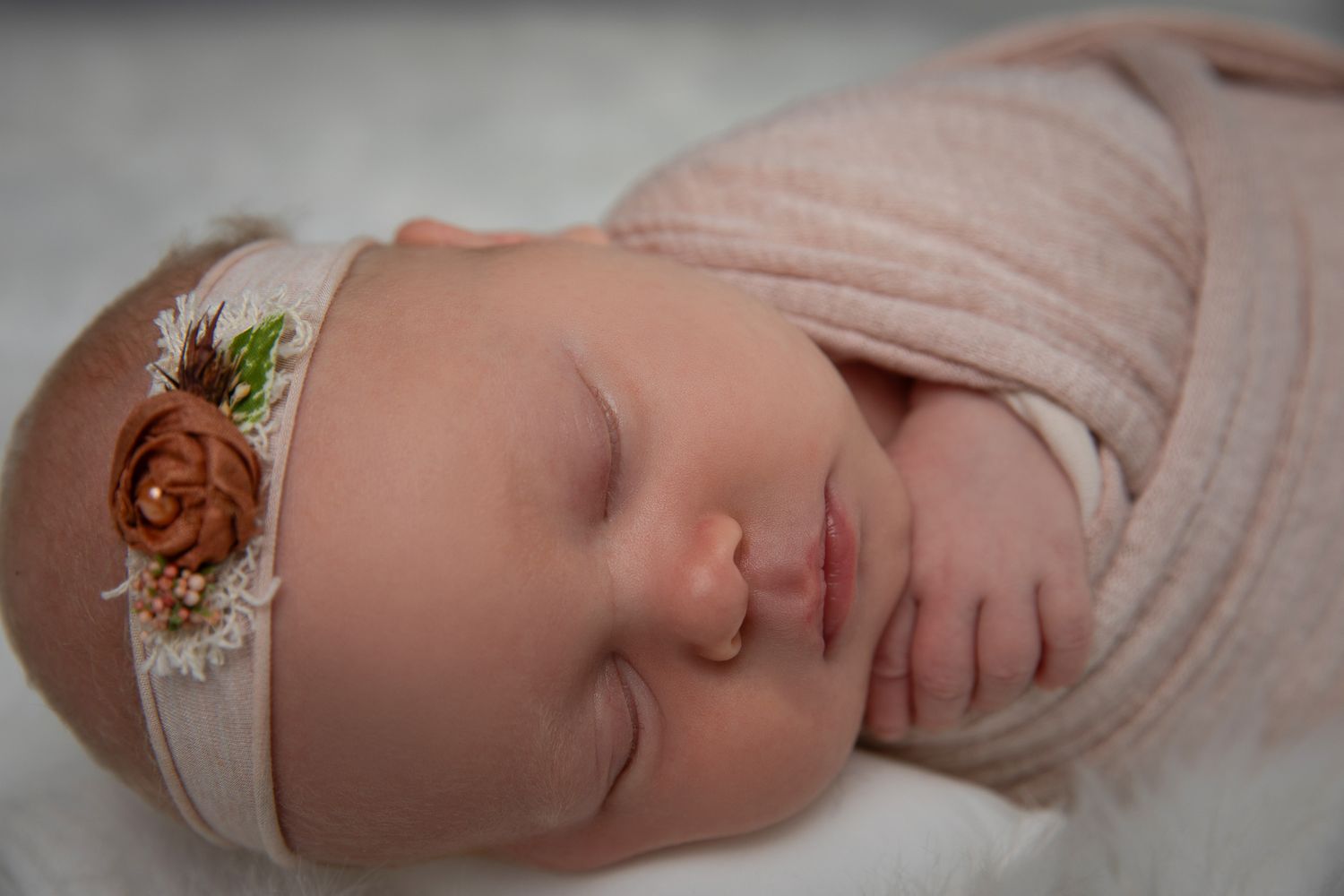 Peaceful newborn baby sleeping wrapped in pink swaddle with floral headband on white background.