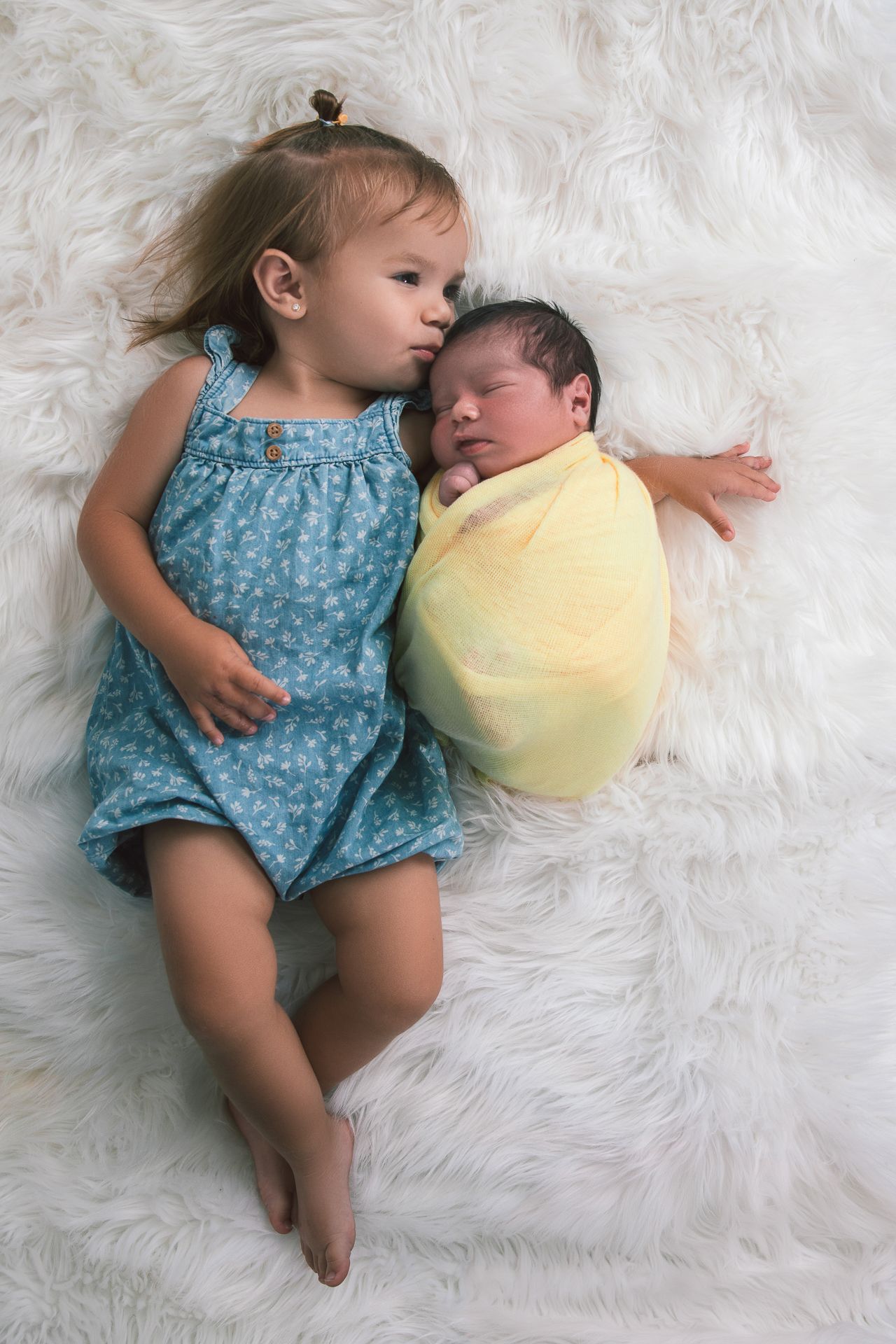 Young girl in blue dress lying beside swaddled newborn baby on white fluffy blanket.