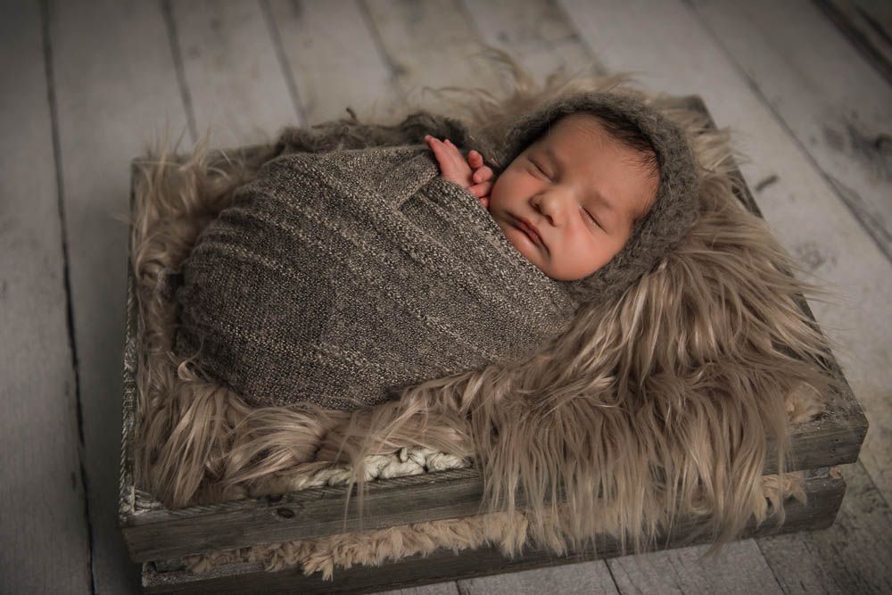 Sleeping newborn baby swaddled in brown knit wrap with bonnet, resting on fluffy fur in rustic wooden crate.