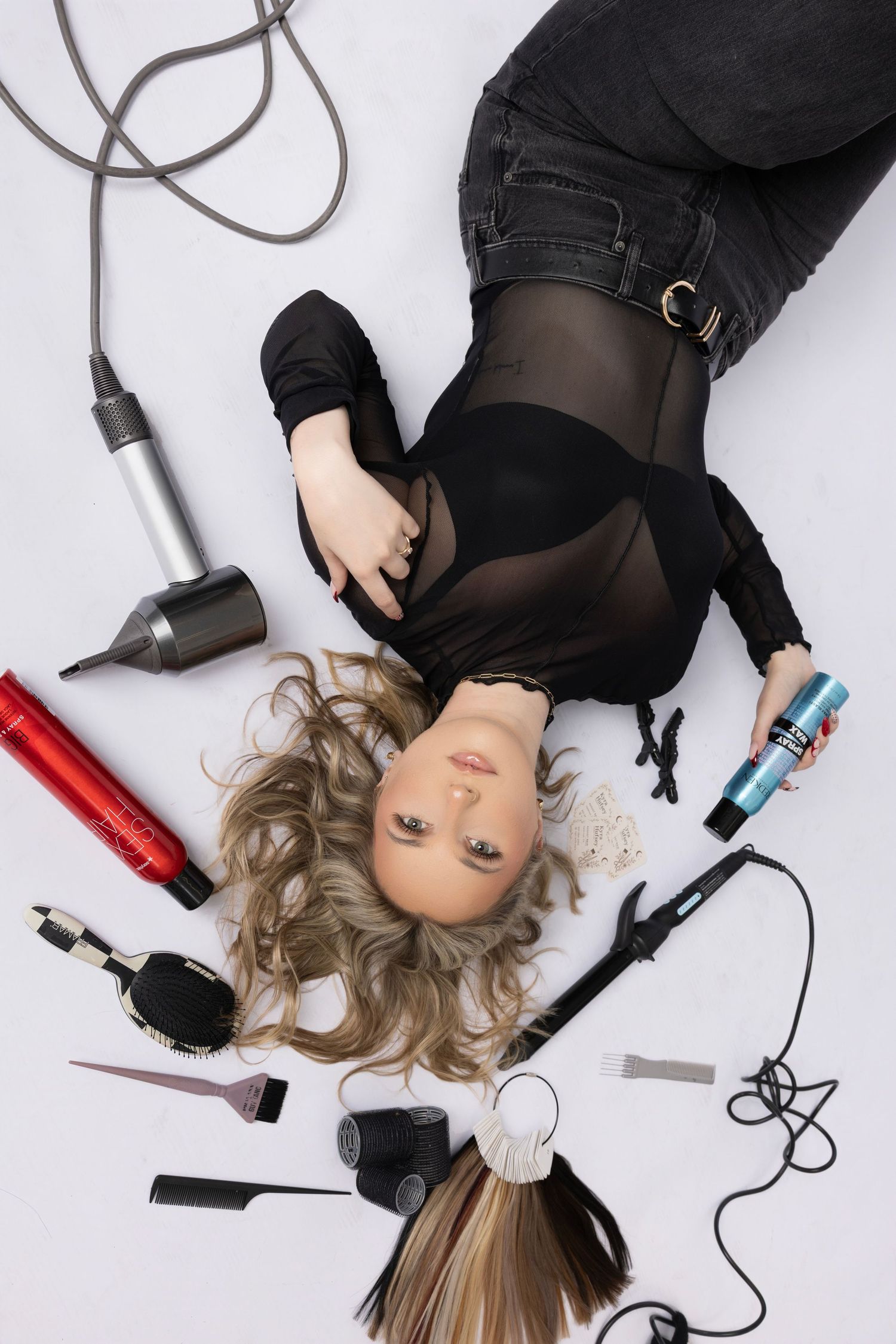 Woman lying on white floor surrounded by hair styling tools including dryer, curling iron, combs, and hair products.
