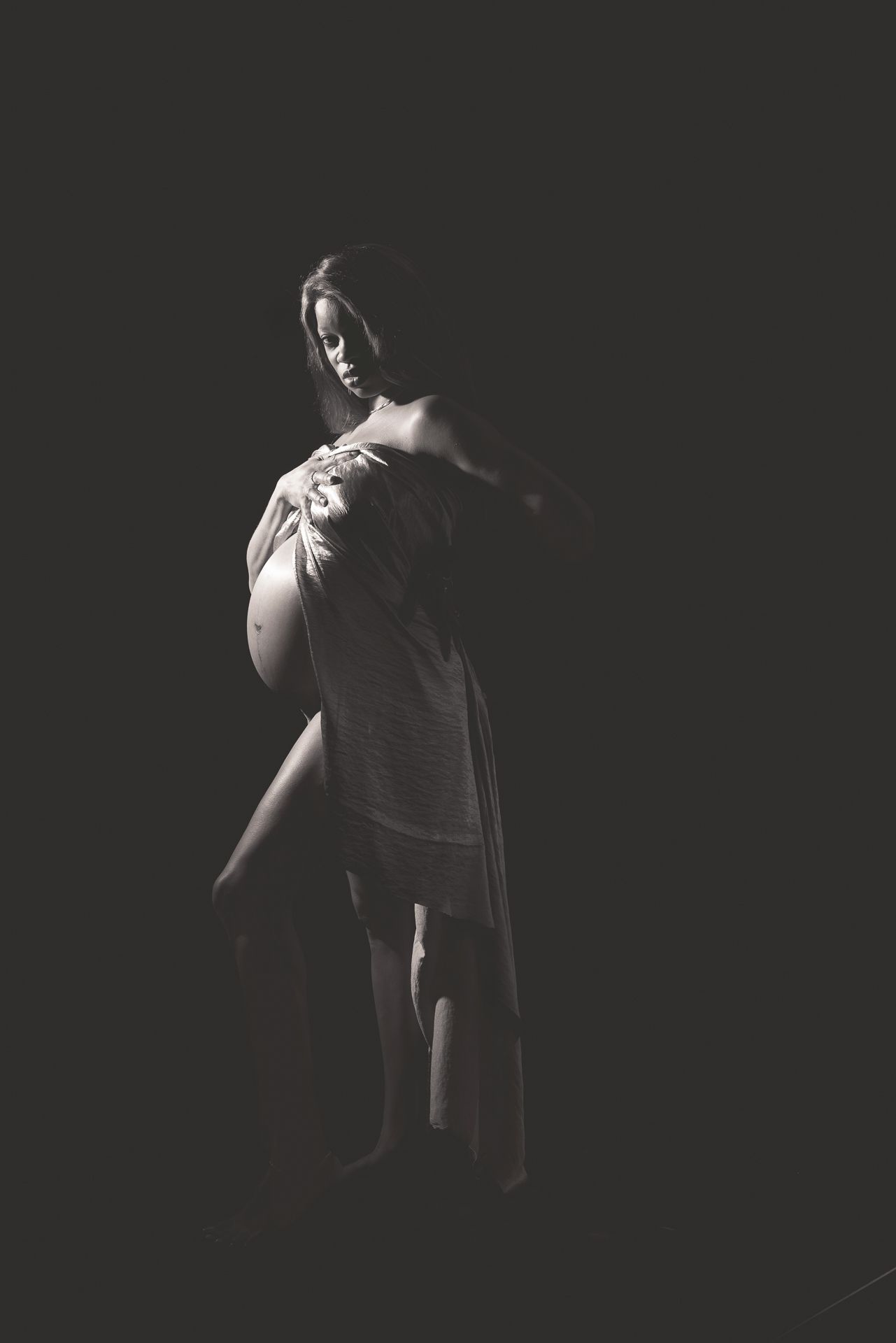 Artistic black and white portrait of pregnant woman draped in fabric, posed in dramatic low-key studio lighting.