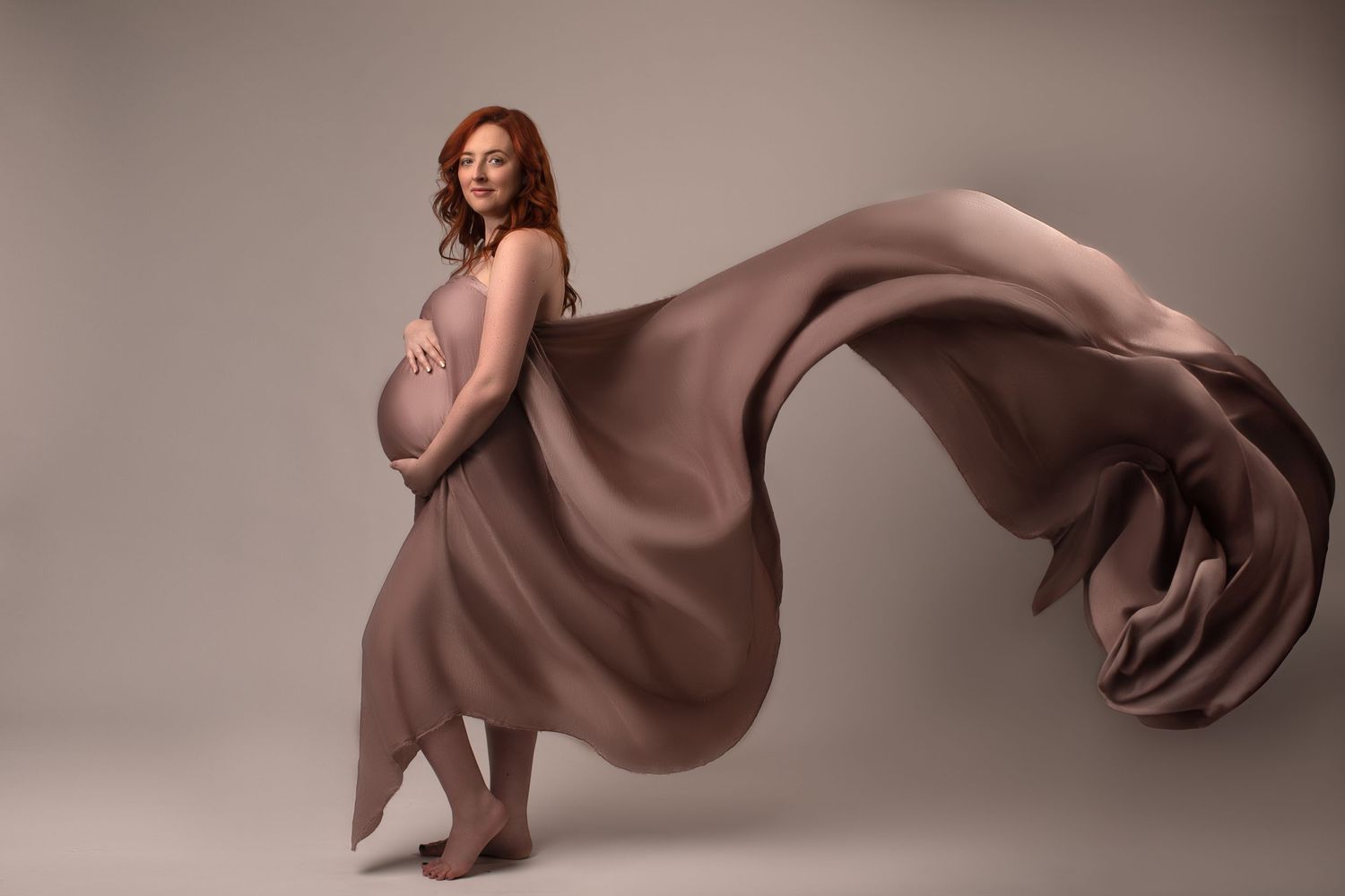 Elegant pregnant woman posed with flowing taupe fabric billowing behind her in a studio maternity shoot.