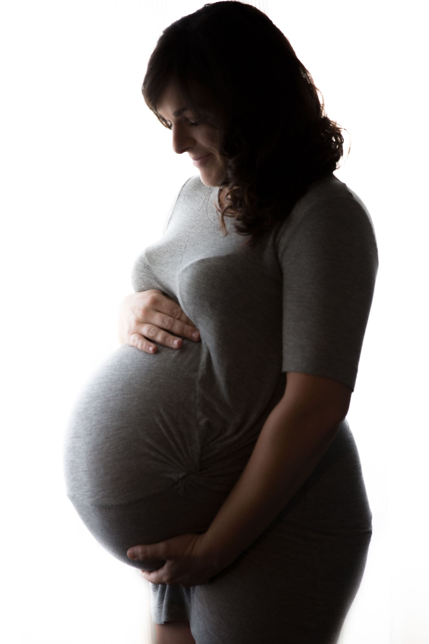 Pregnant woman in gray dress cradling baby bump in dramatic side-lit silhouette portrait.