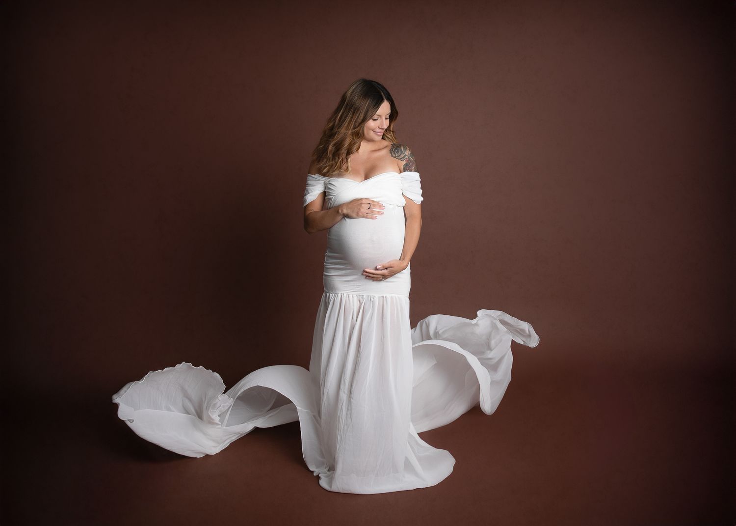 Pregnant woman in flowing white off-shoulder maternity gown with fabric billowing, posing on brown studio backdrop. Bonsall, near Vista,