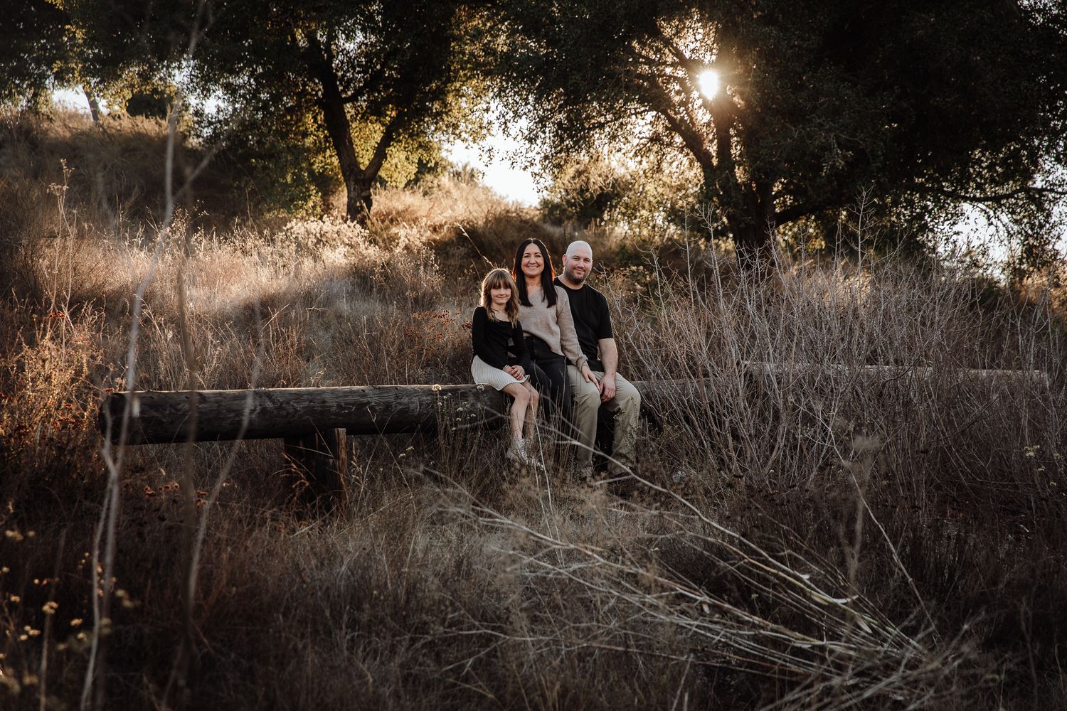 Family of three sitting on log at sunset in golden field surrounded by trees, in Bonsall, near Fallbrook, & Temecula