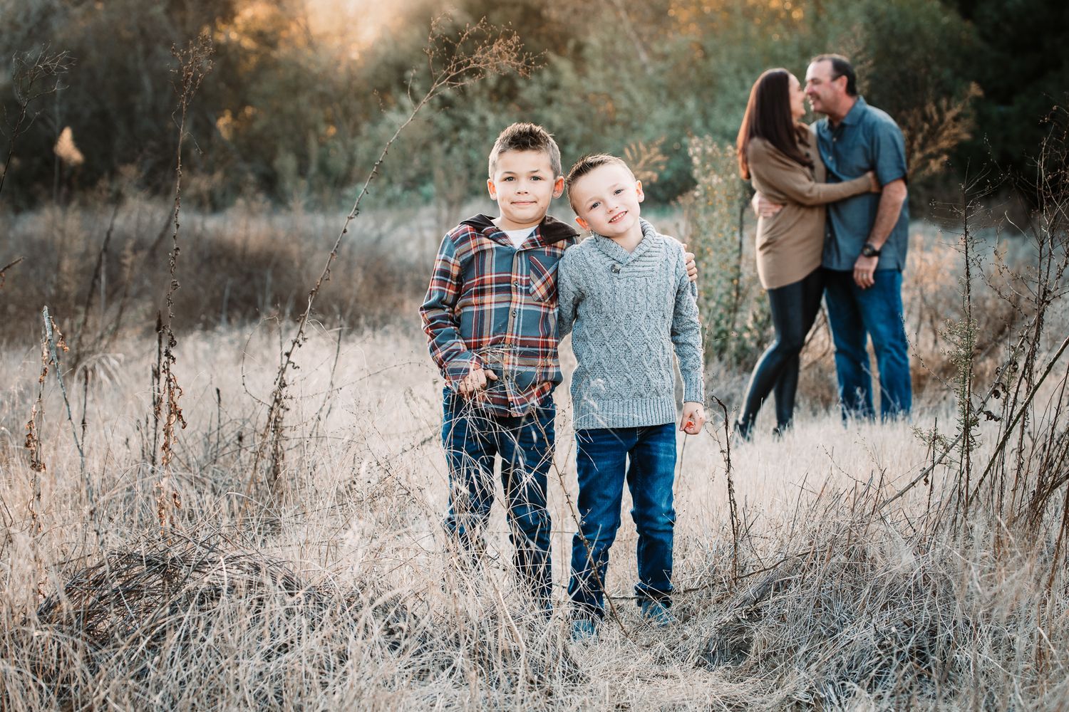 Two young brothers smile together in a field while their parents share a kiss behind them.
