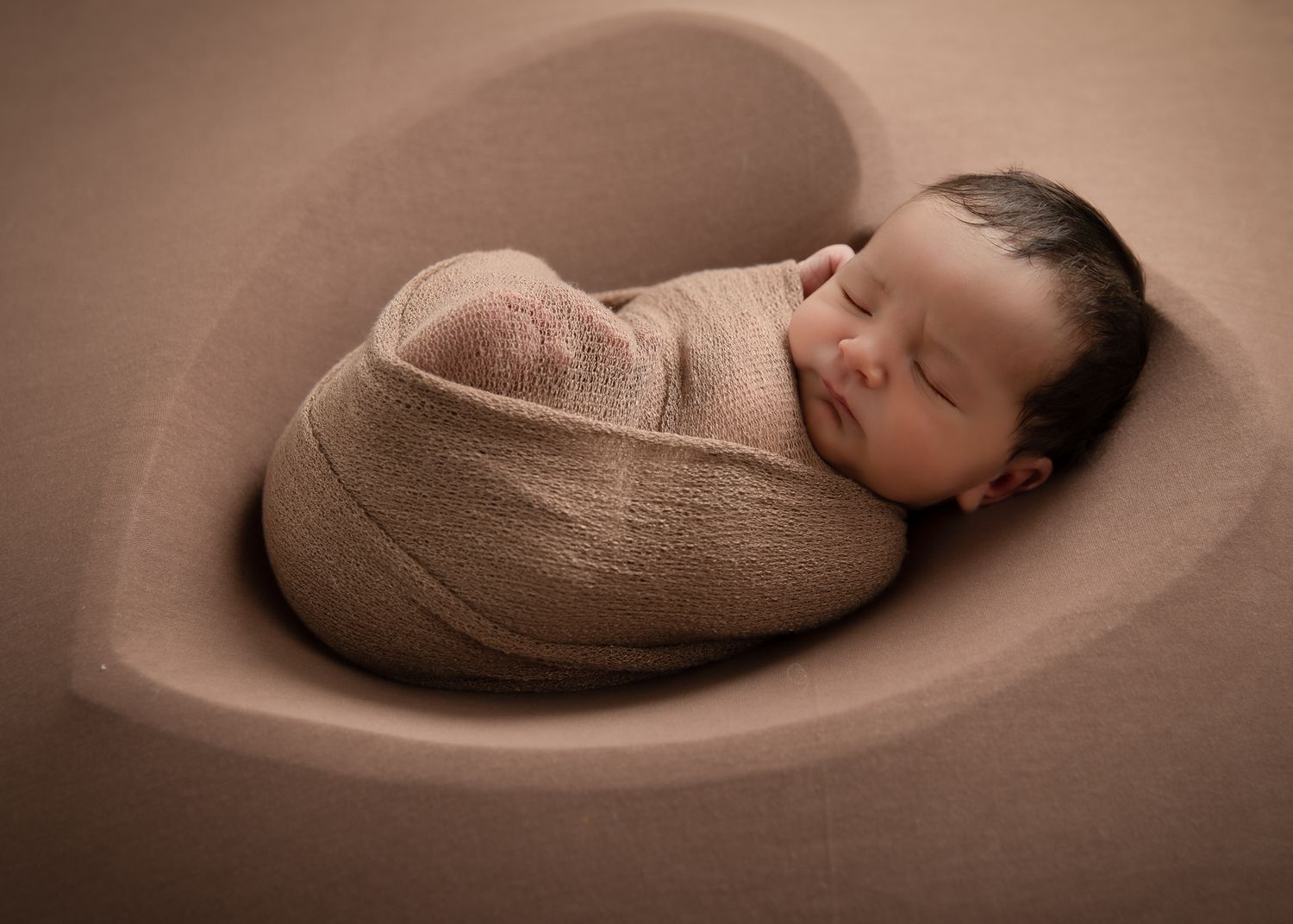 Sleeping dark-haired newborn baby snugly wrapped in a tan swaddle inside a heart-shaped bowl.