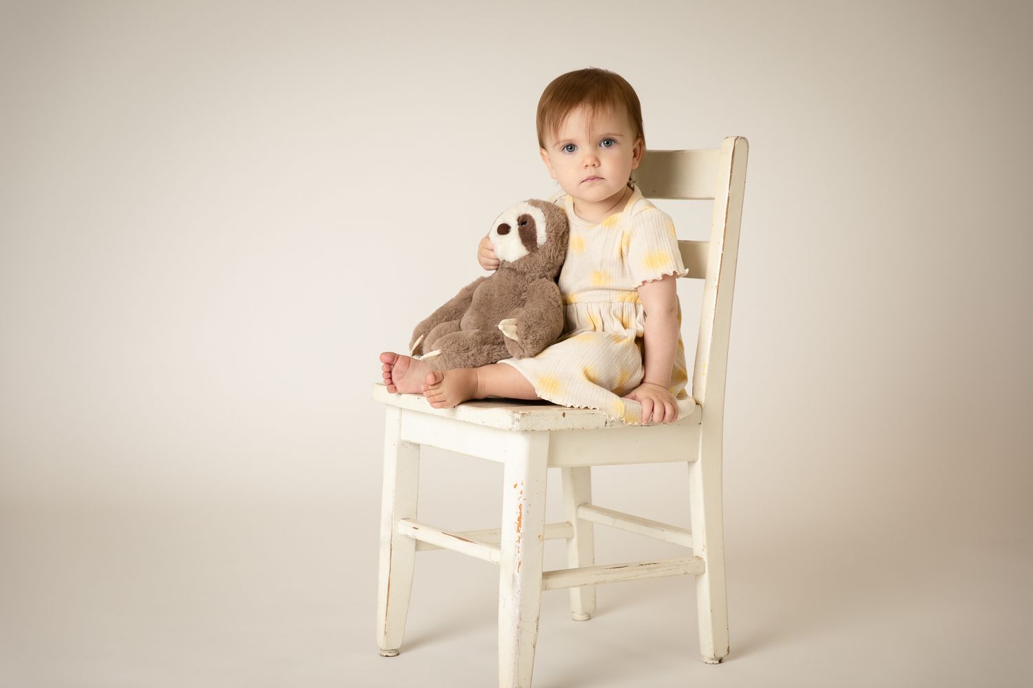 Baby girl in yellow dress sitting on white chair with teddy bear.