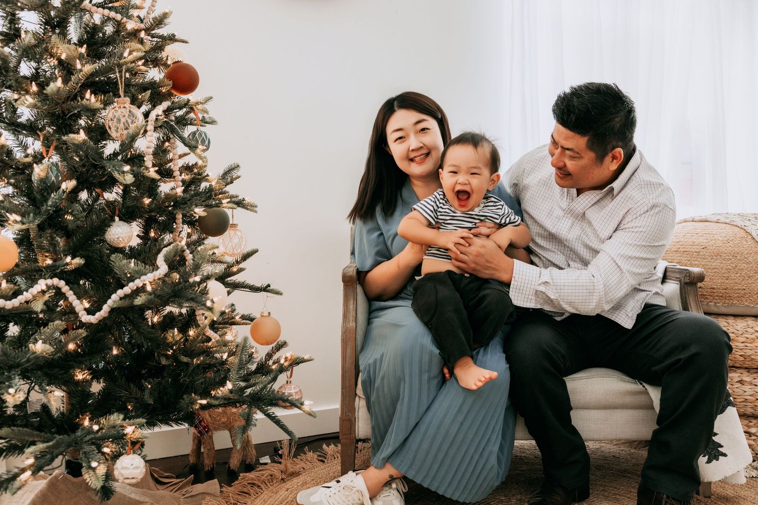 Happy Asian family with baby sitting near decorated Christmas tree in cozy white living room holiday portrait.