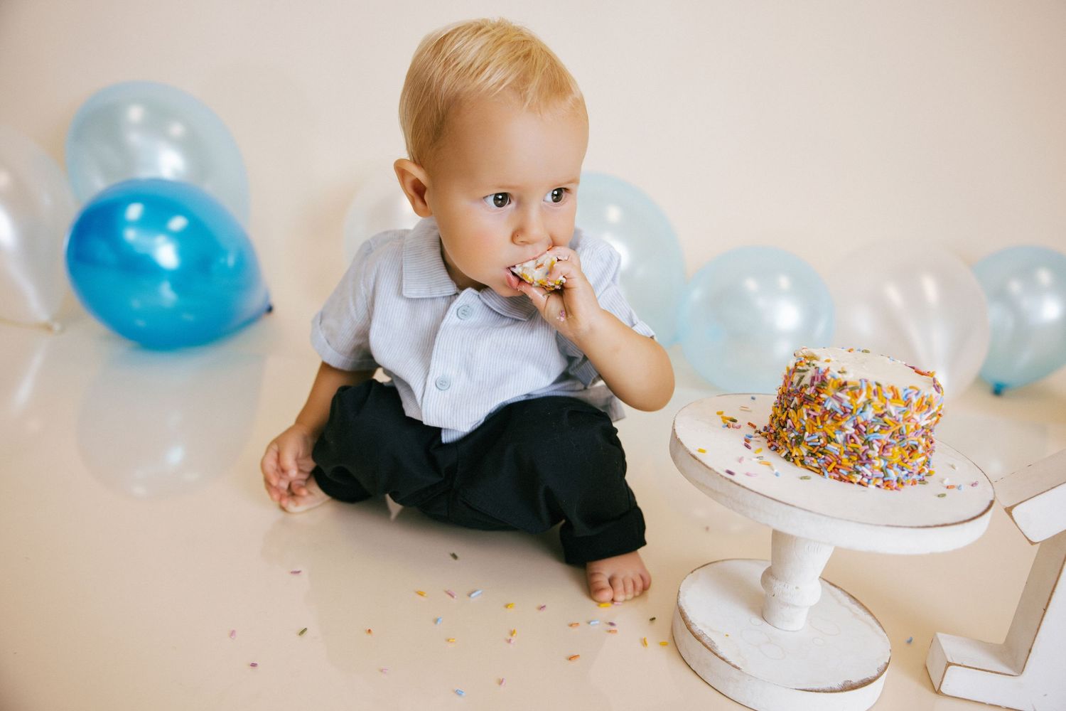 Baby boy in blue shirt eating birthday cake with colorful sprinkles, surrounded by blue balloons at first birthday party.