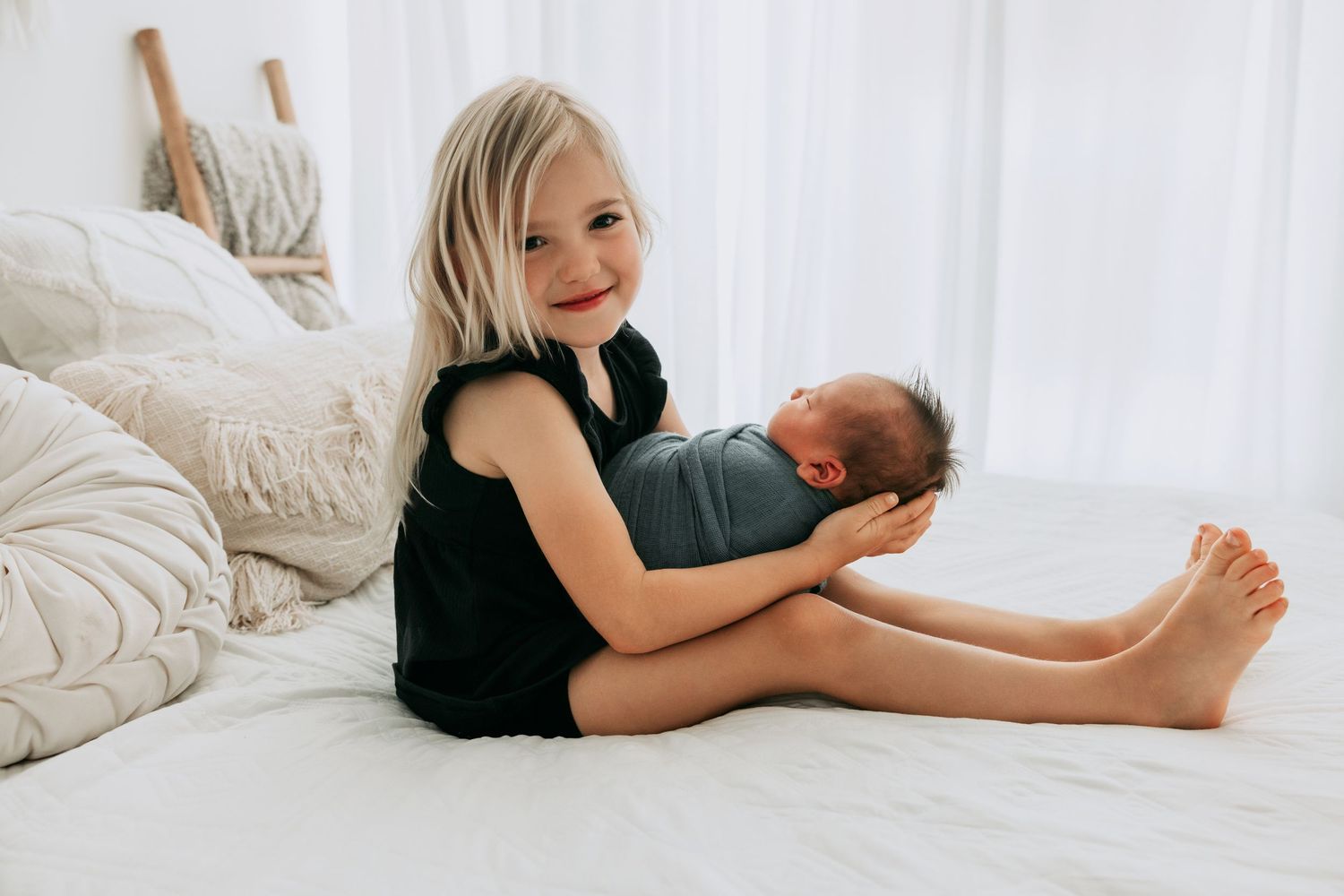 Smiling toddler girl sitting on white bed holding sleeping newborn baby sibling during professional photo session.