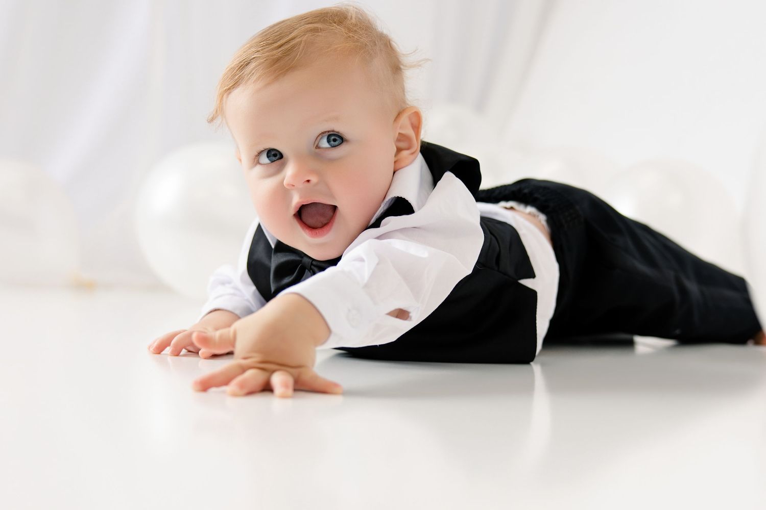 Adorable baby in a black tuxedo outfit crawling on a white floor, smiling at the camera.