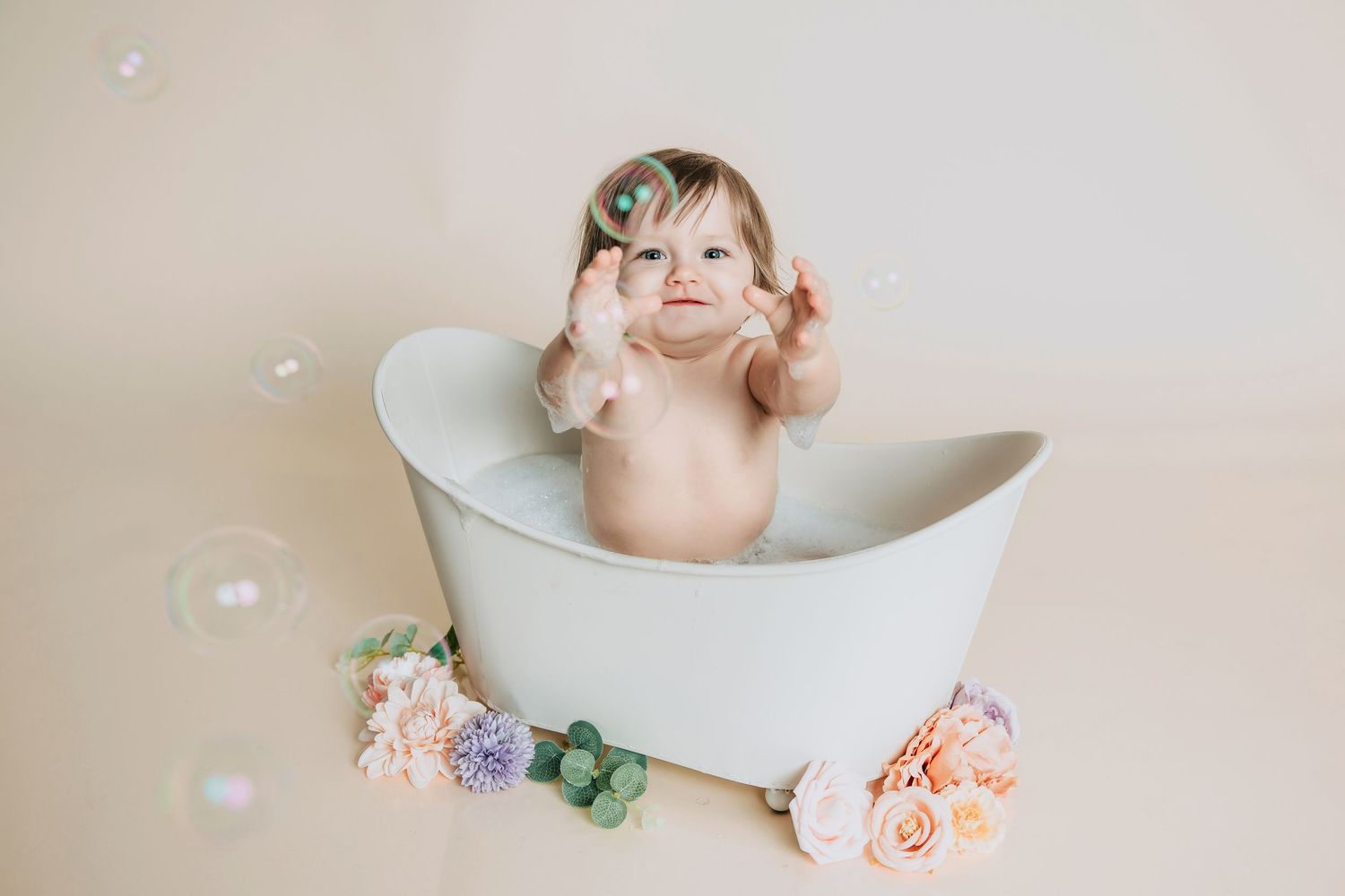 Baby girl sitting in a white mini bathtub playing with bubbles, surrounded by pastel flowers on a beige background.