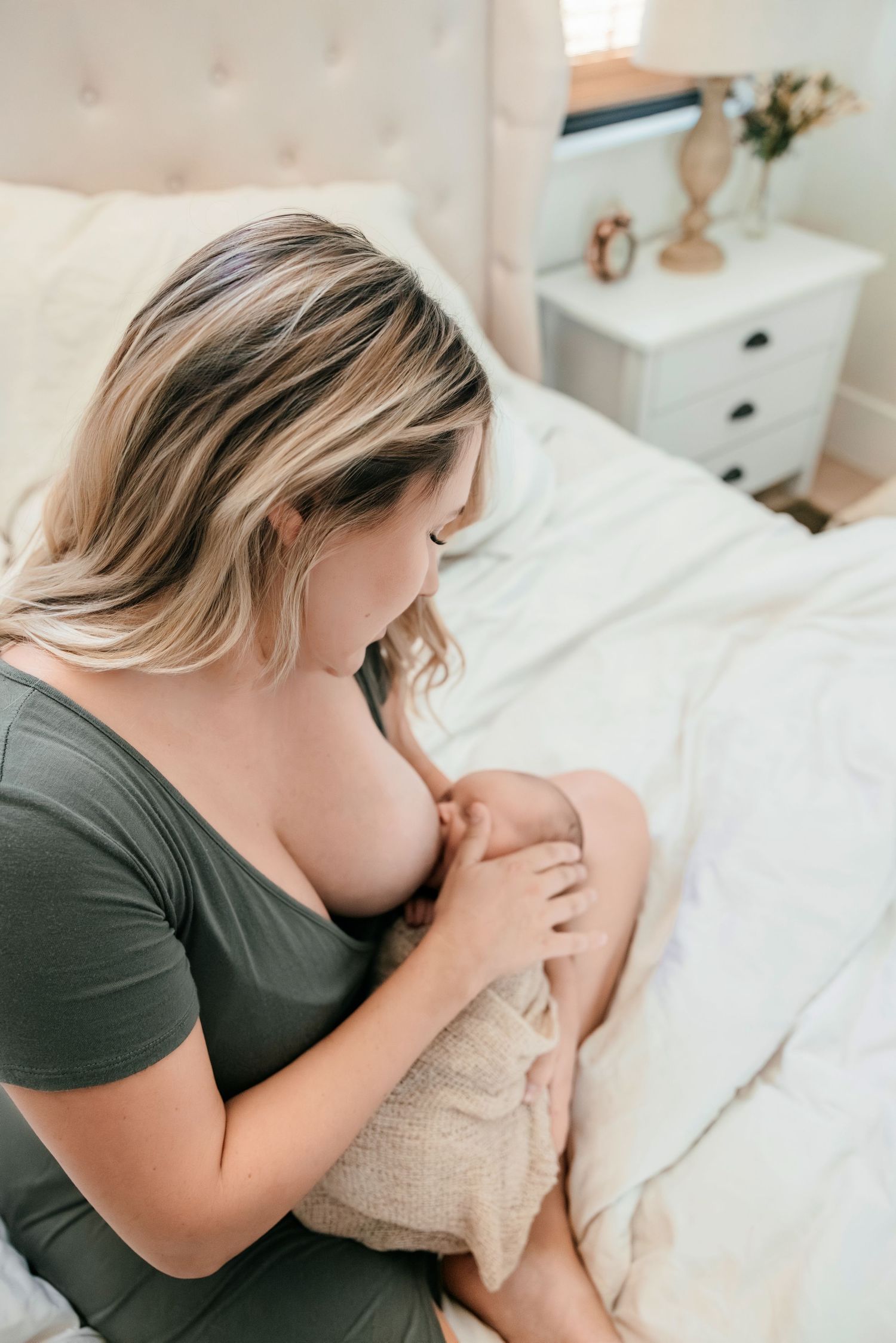 A mother breastfeeding her newborn baby while sitting on a bed in a cozy bedroom setting.