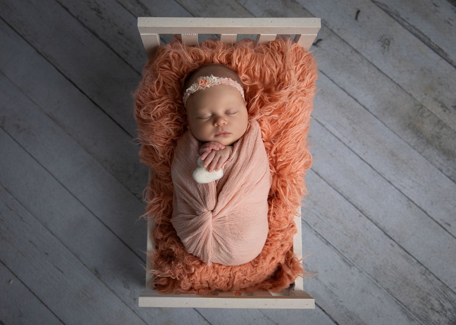 Newborn baby wrapped in peach swaddle with floral headband sleeping on fluffy orange fur in a small wooden crib.