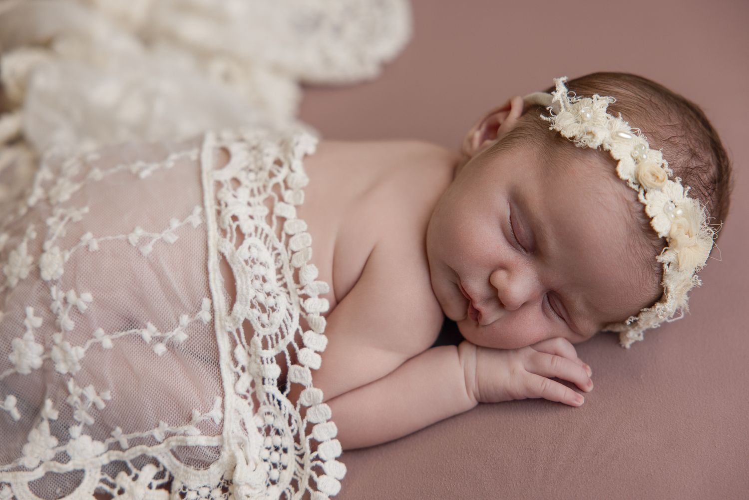 Sleeping newborn baby wrapped in white lace with a floral headband on a mauve background.