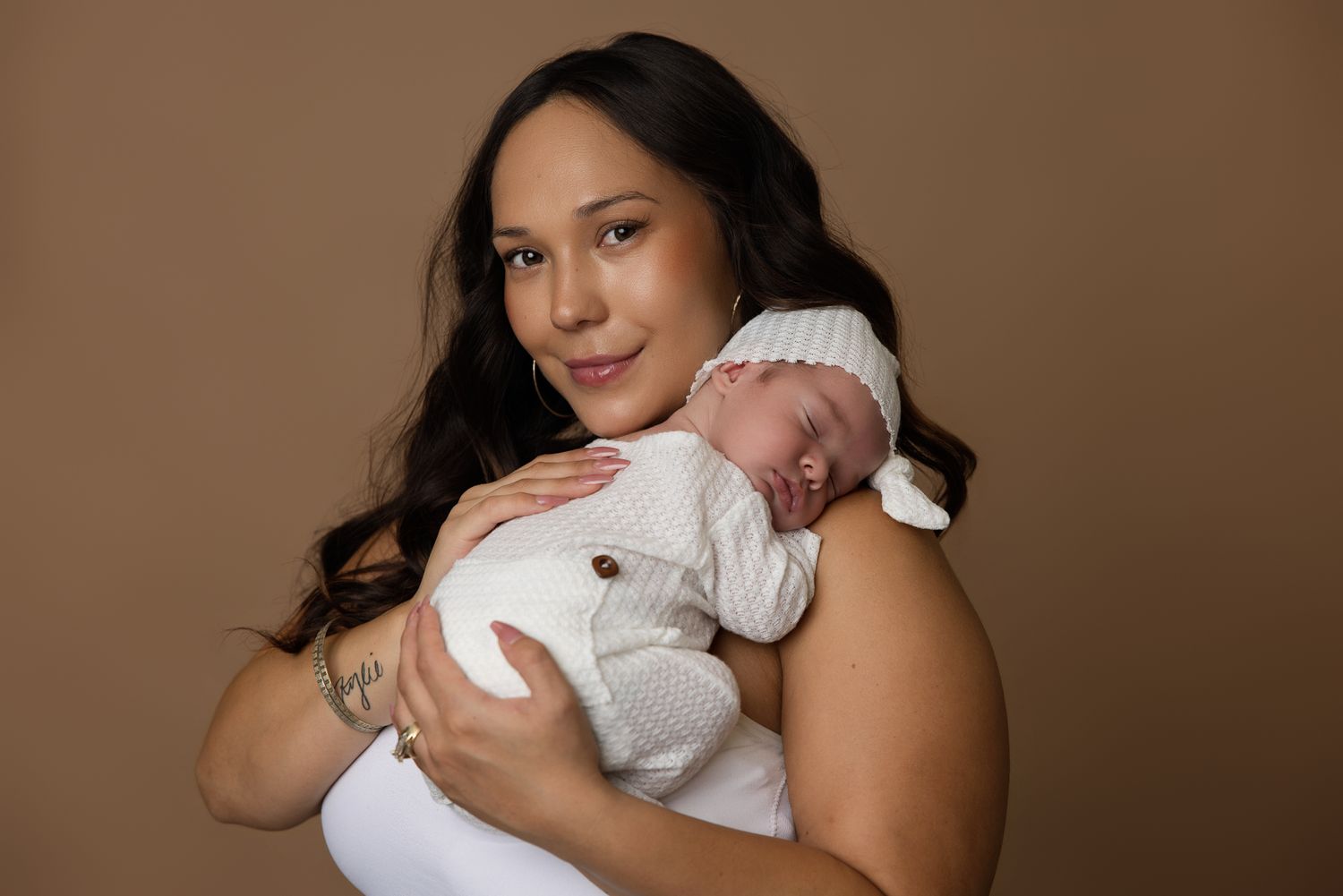 A mother tenderly holds her sleeping newborn baby dressed in white against a warm brown studio background.