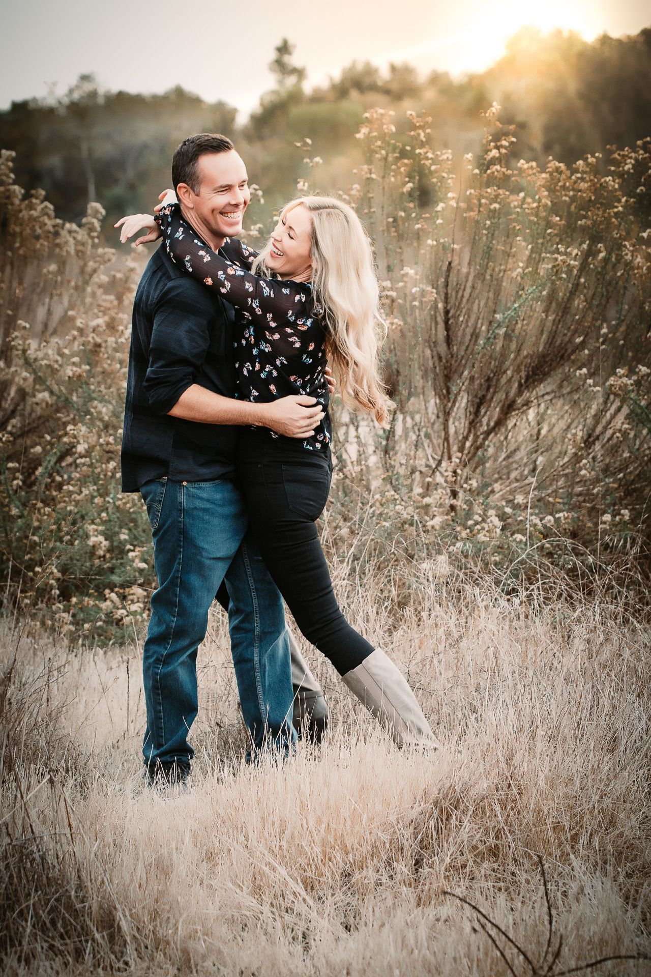 A happy couple embraces and smiles at each other in a golden sunlit field at sunset.
