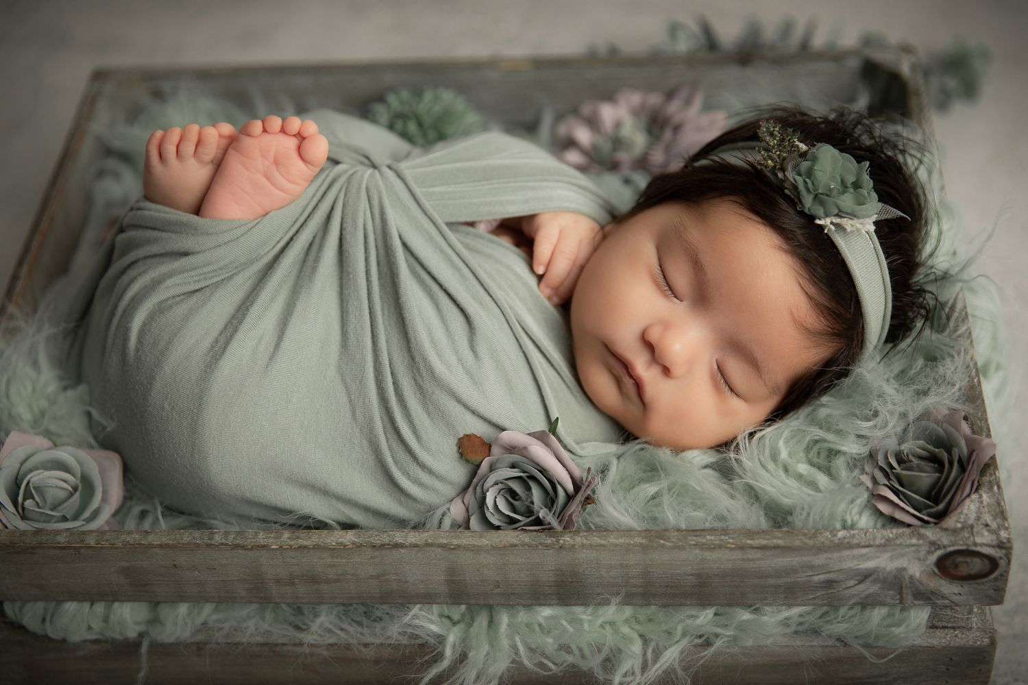 Sleeping newborn girl swaddled in sage green wrap rests in a wooden box with rose decorations in Bonsall, near Carlsbad