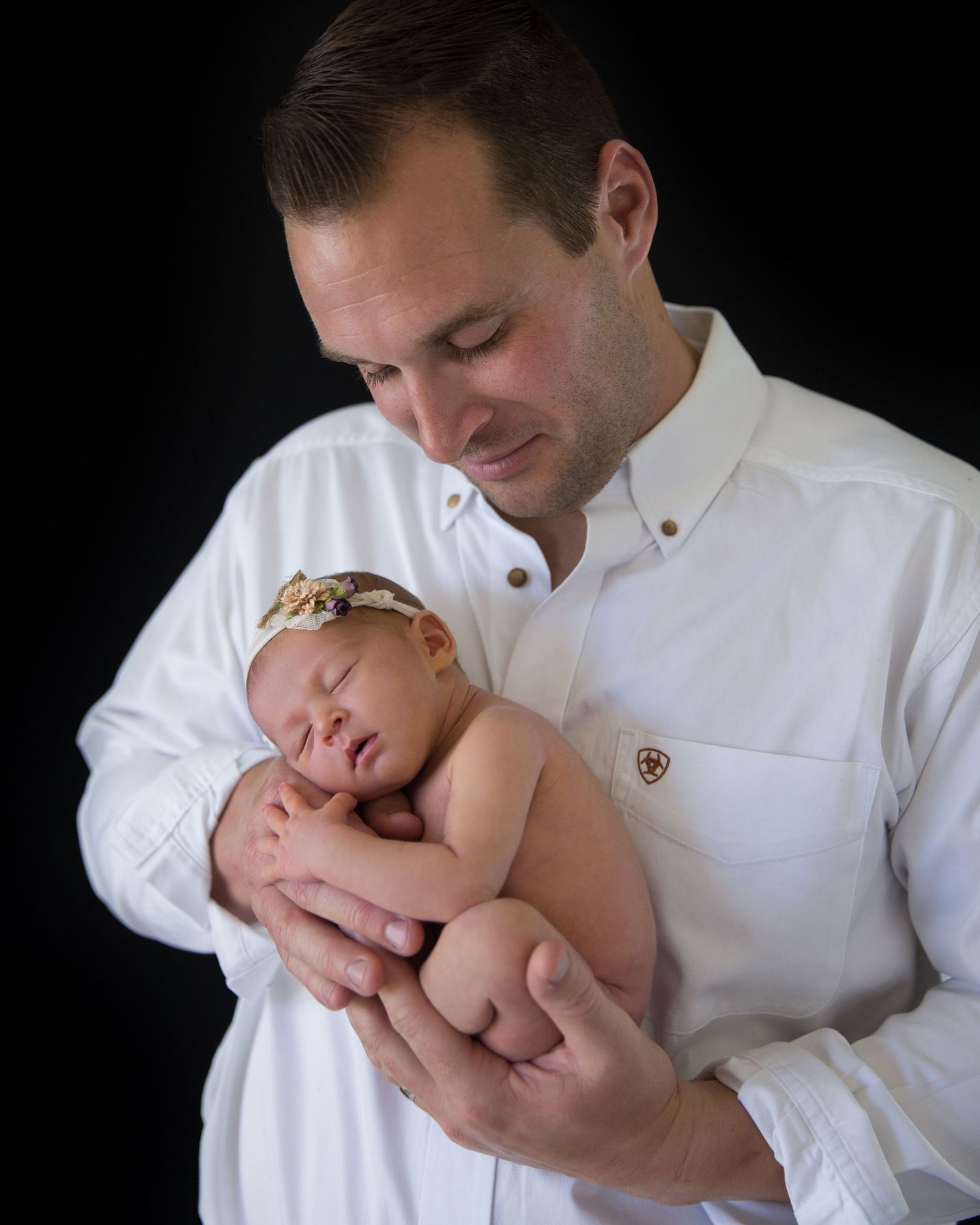 A man in a white shirt tenderly holds a sleeping newborn baby girl wearing a floral headband against a dark background.