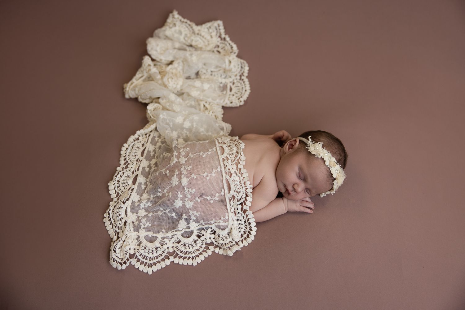 Newborn baby sleeping peacefully on lace fabric with floral headband on brown background.