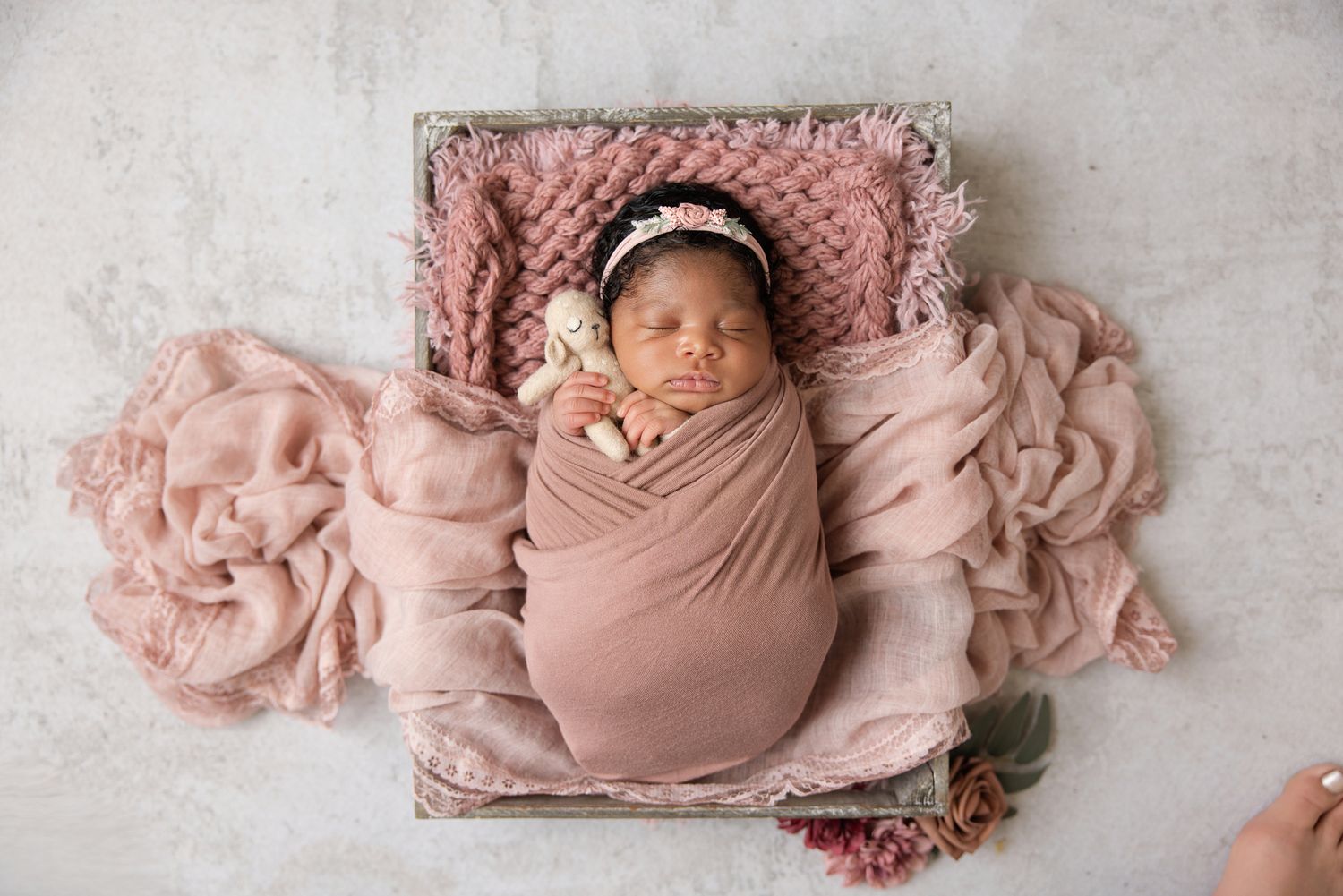 Newborn baby girl swaddled in pink wrap, sleeping peacefully in a wooden box with floral headband.