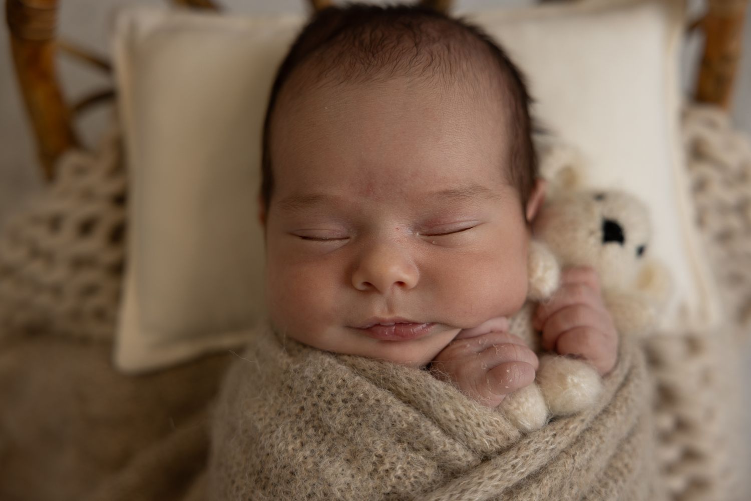 Peaceful newborn baby swaddled in a soft beige knit wrap, sleeping beside a tiny teddy bear on a cozy blanket.