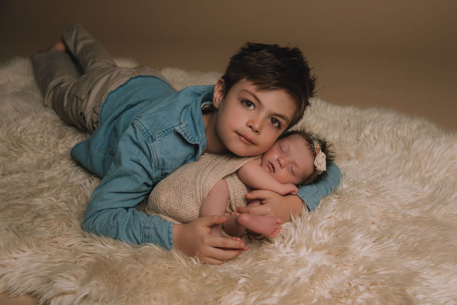 Older boy in denim shirt lying on fluffy white rug tenderly holding sleeping newborn baby with floral headband.