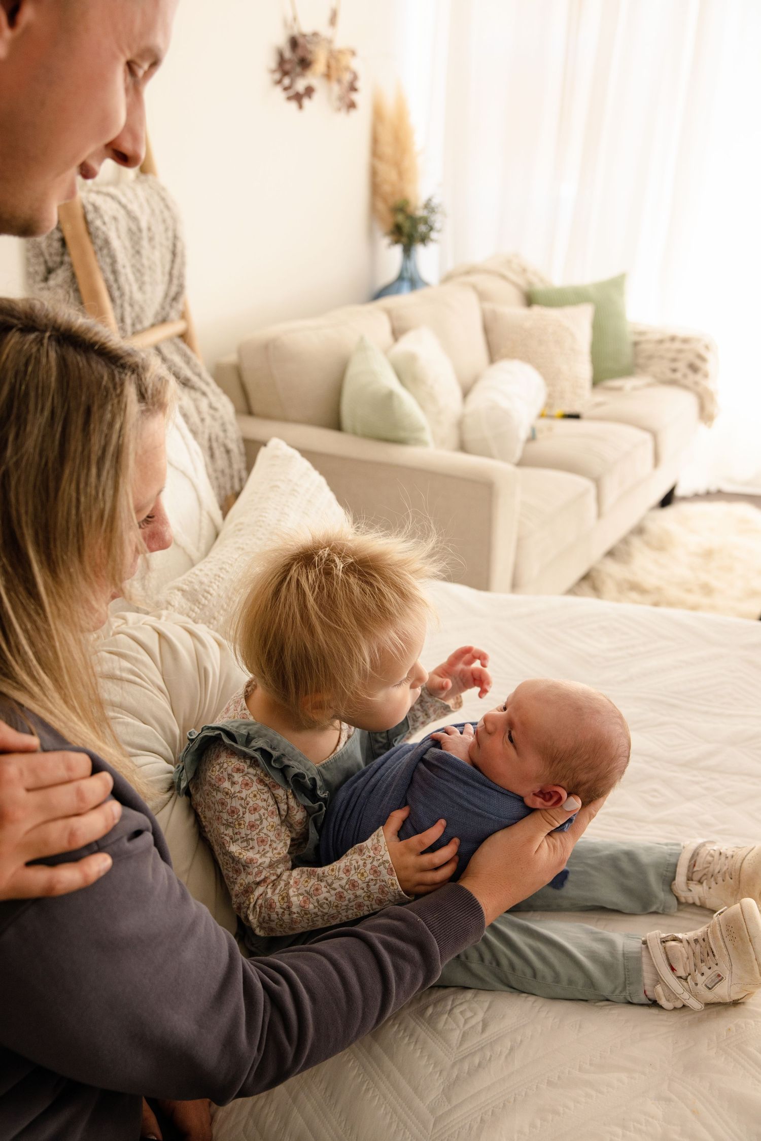 A young family sits on a bed, with a toddler gently touching a swaddled newborn baby held by the mother.