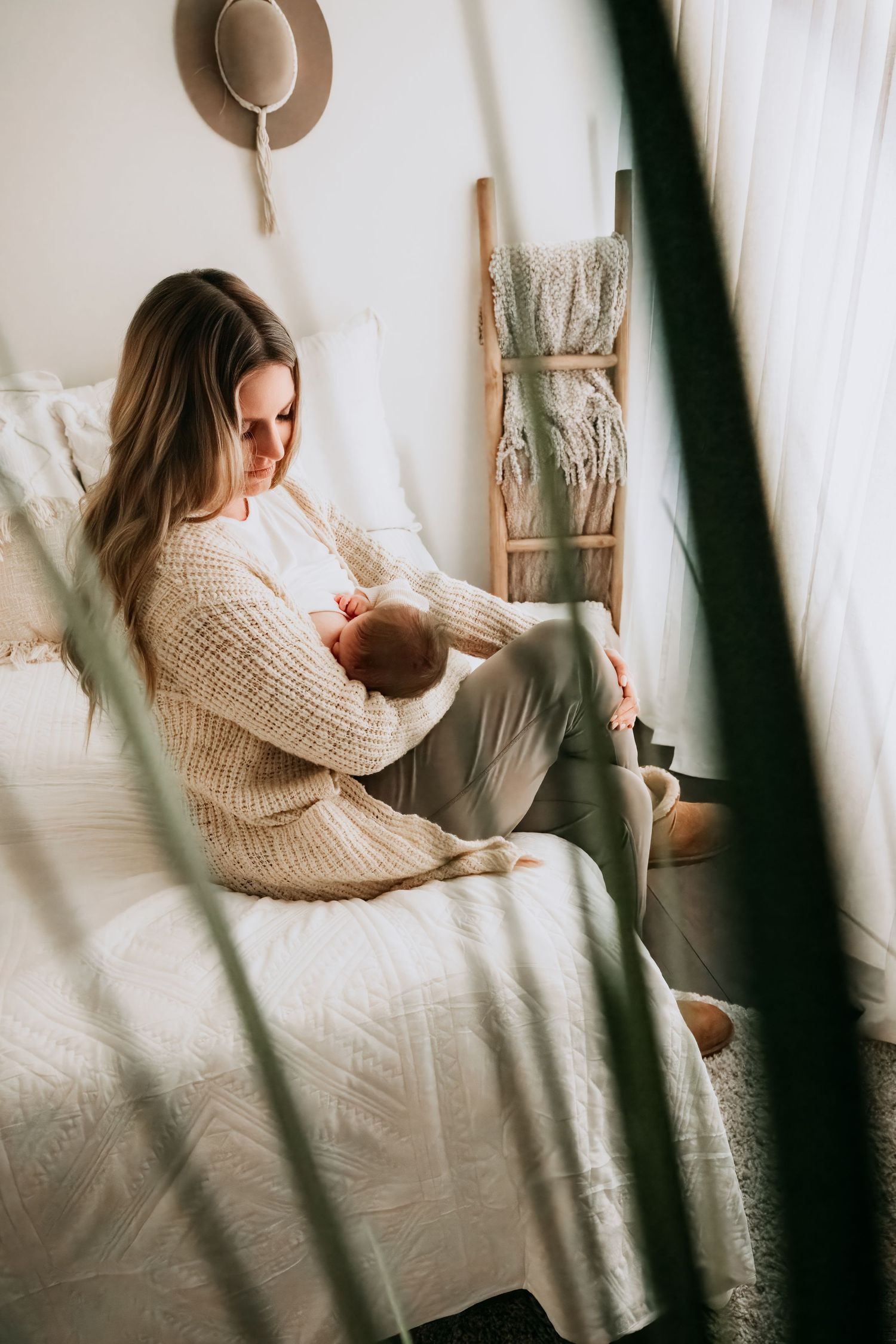 A mother in a cream knit sweater sits on a white bed, tenderly cradling her newborn baby in a bright, cozy room.