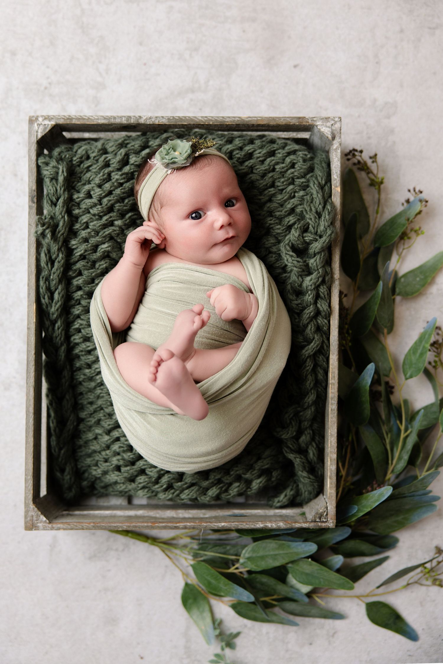 Newborn baby swaddled in sage green wrap lying in wooden crate with knit blanket and eucalyptus branches.