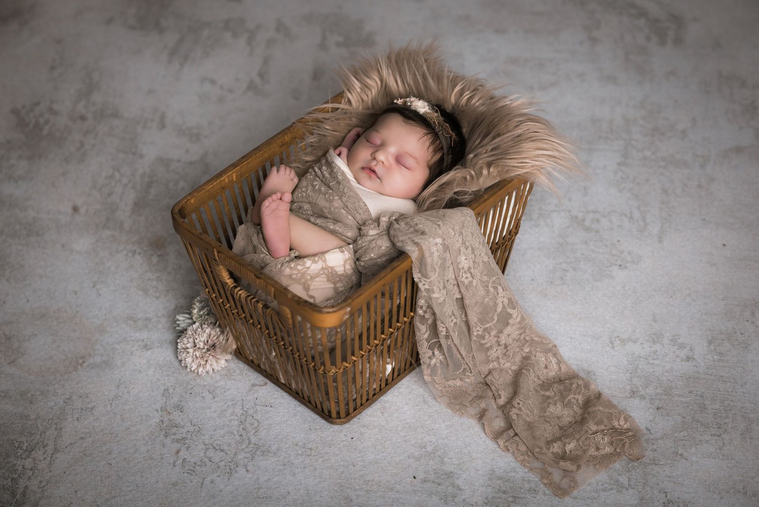 Newborn baby sleeping peacefully in a rustic wicker basket with soft beige lace wrap and floral headband on gray backdrop.