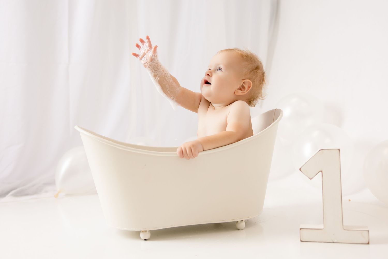 Happy baby sitting in a white mini bathtub reaching up, celebrating their first birthday in a bright white studio.