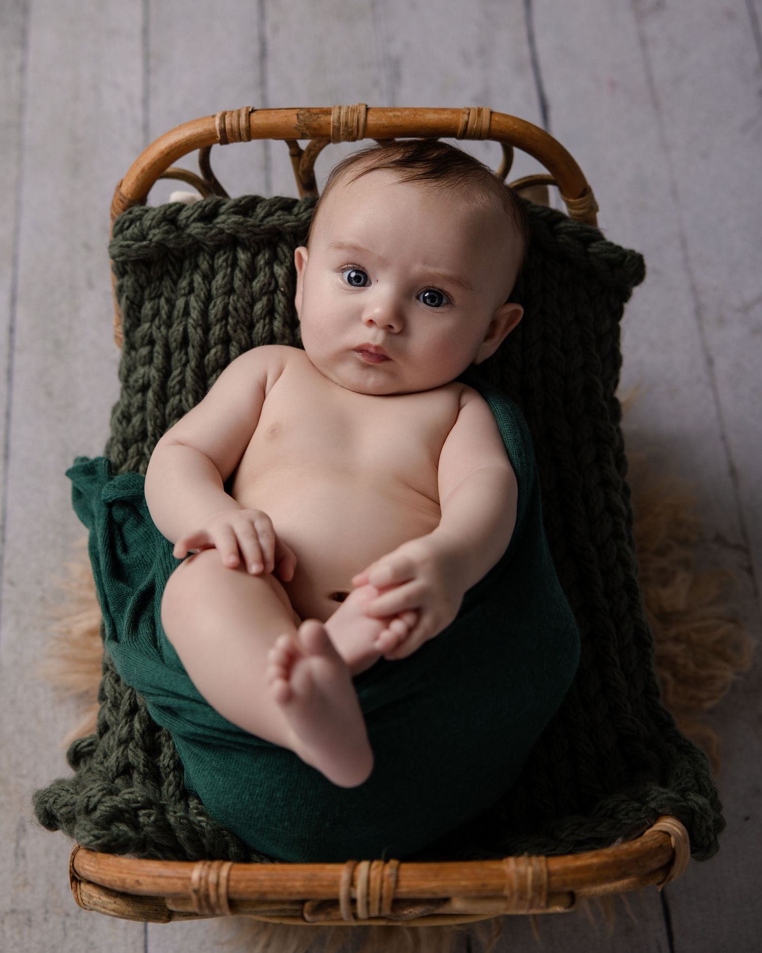 A baby rests in a small rattan bed with a green knit blanket, looking up with bright eyes.