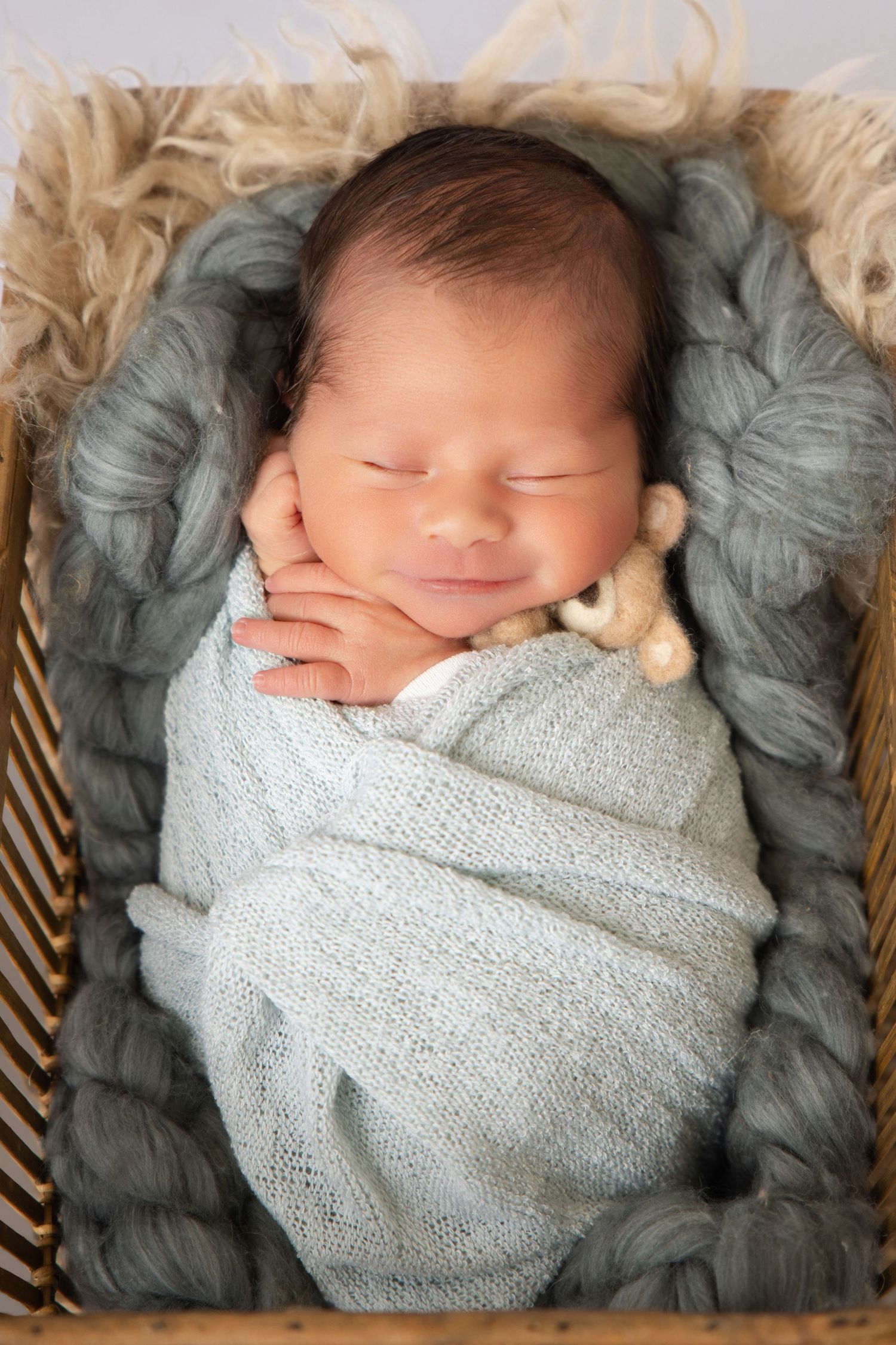 Smiling newborn baby swaddled in soft blue wrap, sleeping peacefully in a wicker basket with a tiny teddy bear.
