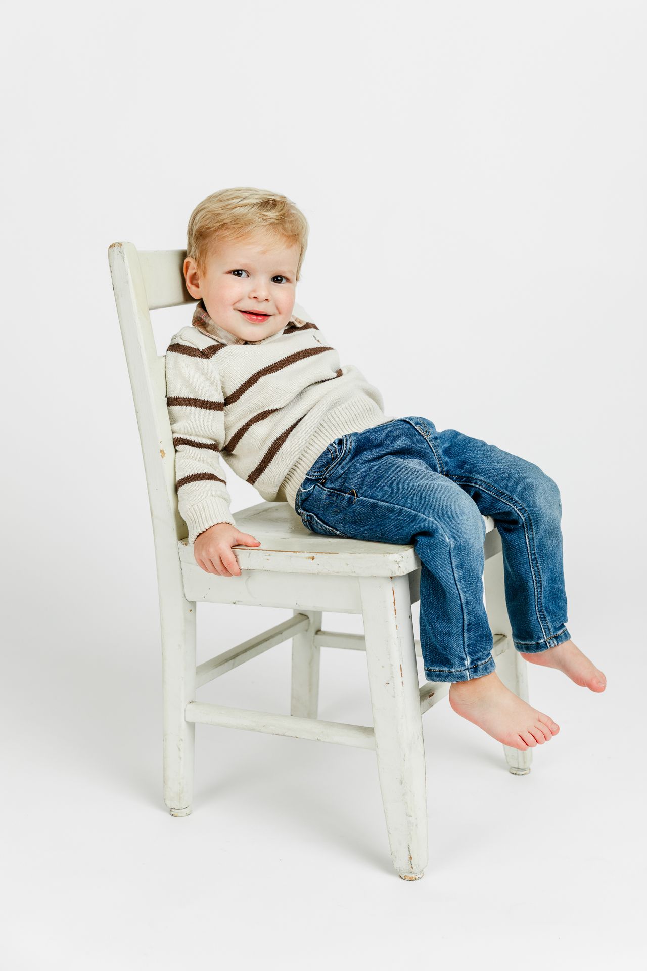 Cute toddler in striped sweater and jeans sitting barefoot on a white wooden chair, smiling.