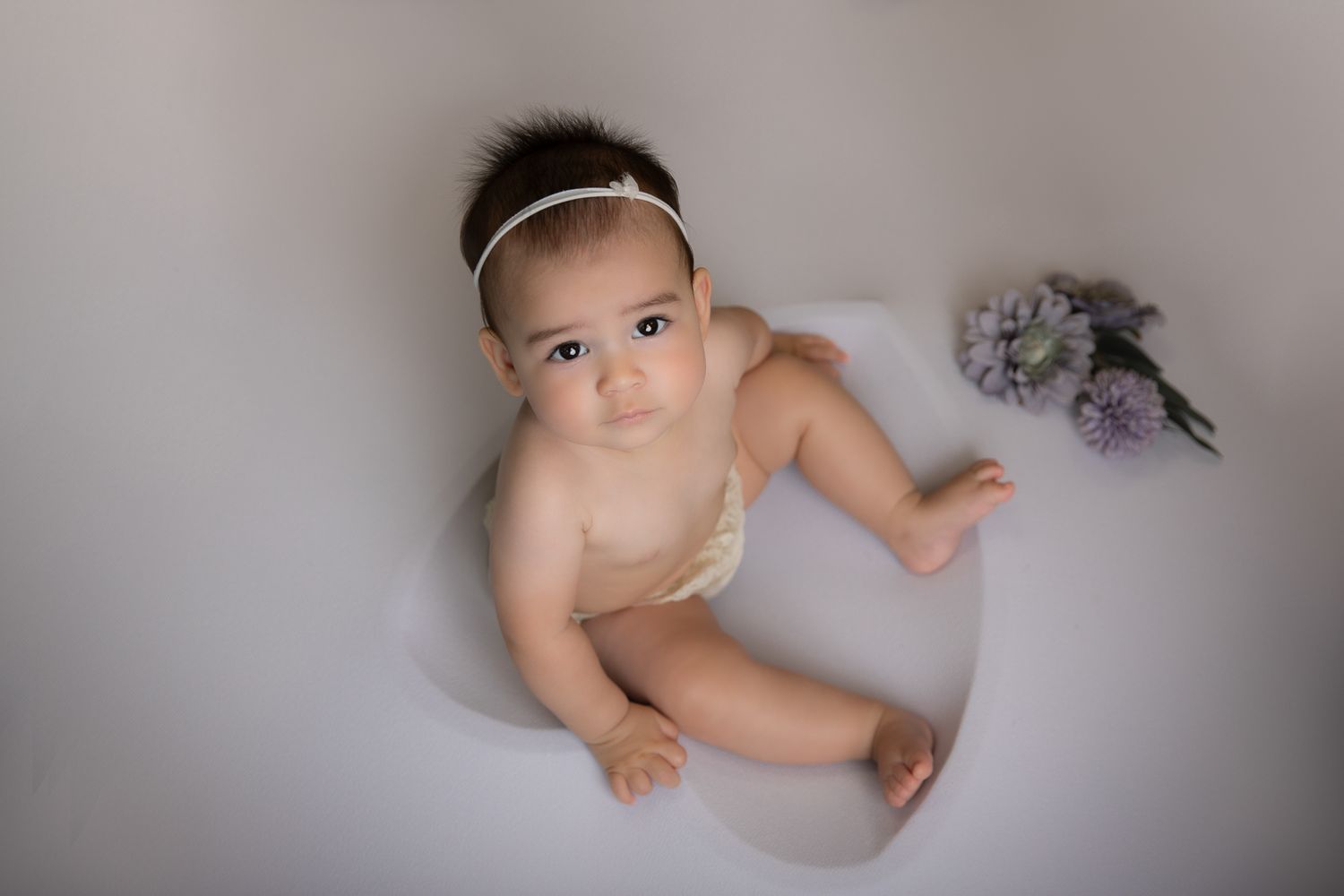 Adorable baby sitting in milk bath with purple flowers.