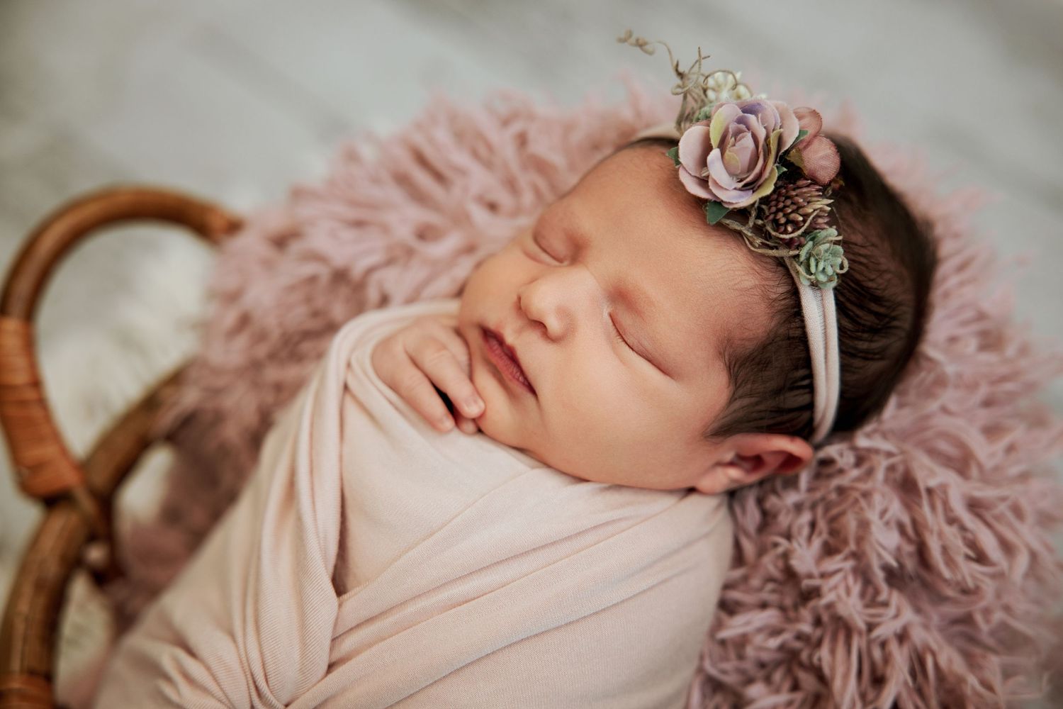 Sleeping newborn baby girl wrapped in pink swaddle with floral headband resting in a basket.