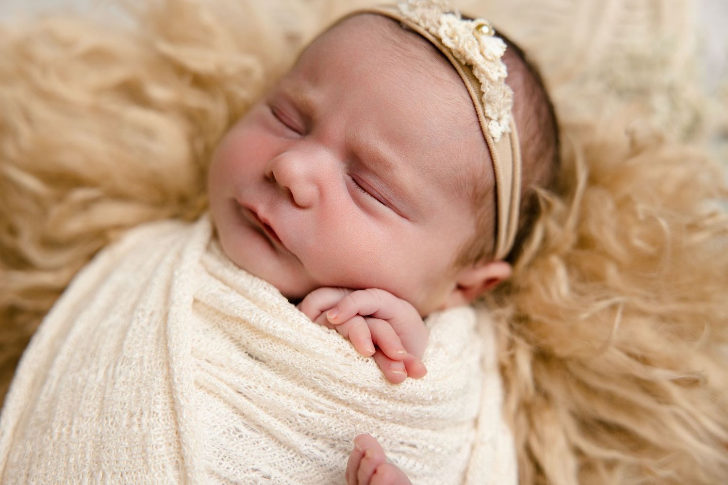 Sleeping newborn baby girl swaddled in cream wrap with gold headband on fluffy fur backdrop.