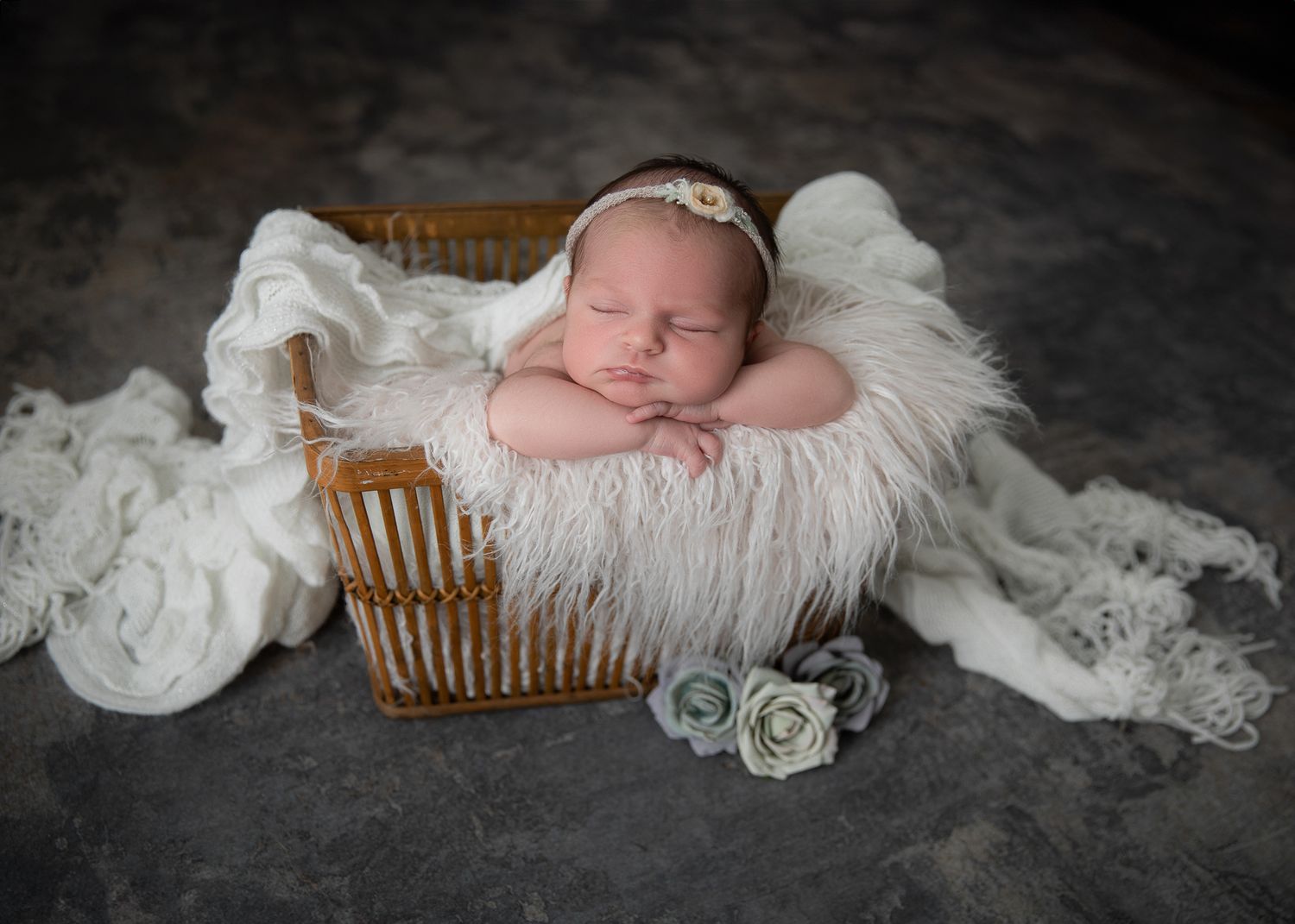 Sleeping newborn baby in a wicker basket with white fur, wearing a floral headband, surrounded by soft roses.