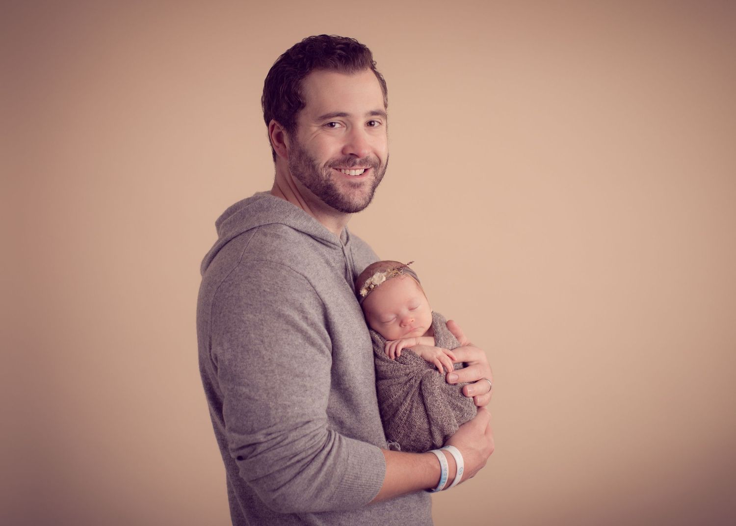 A smiling father holds his sleeping newborn baby wrapped in a brown swaddle against a beige studio background.