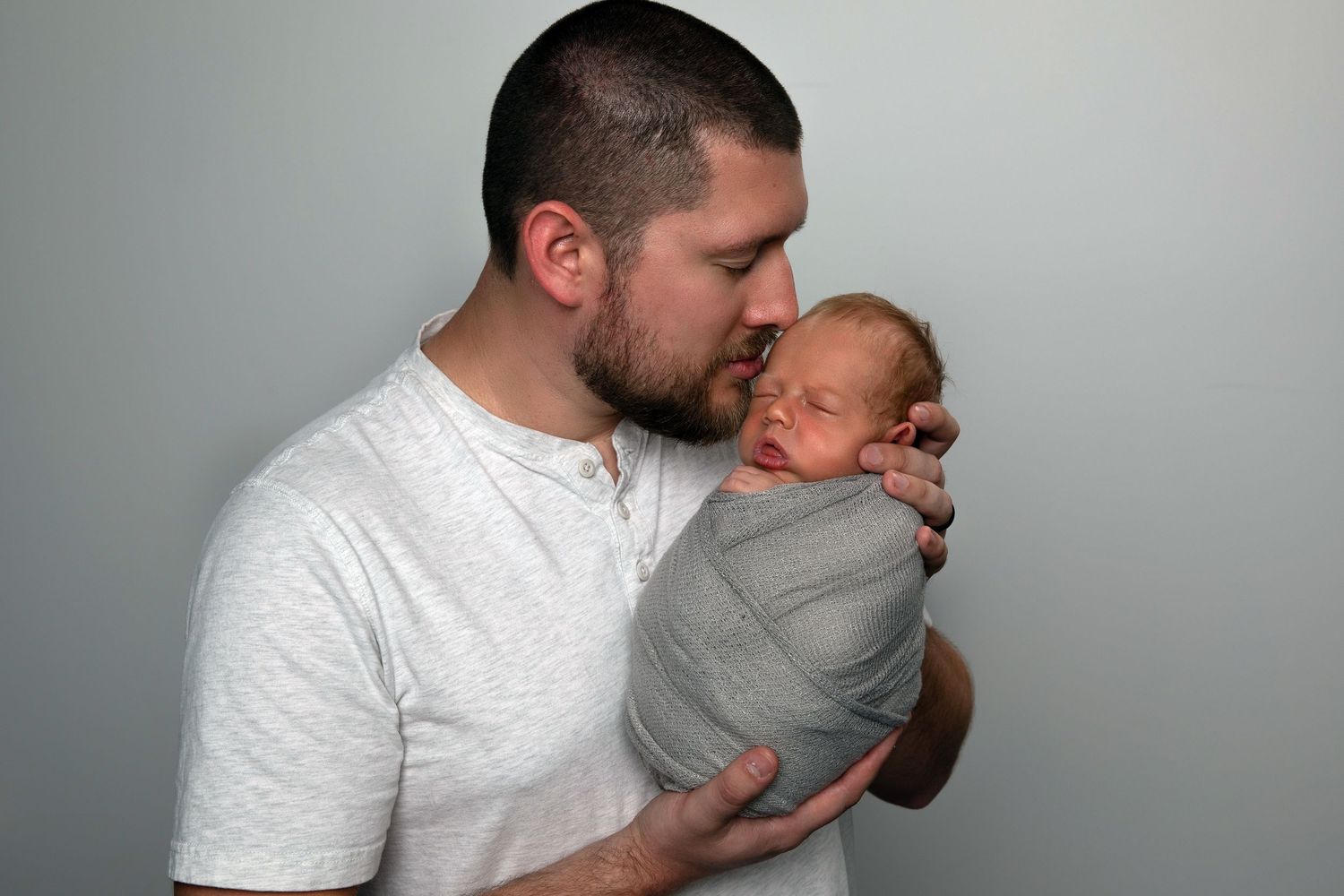 A father tenderly kisses his swaddled newborn baby wrapped in gray cloth against a light background.