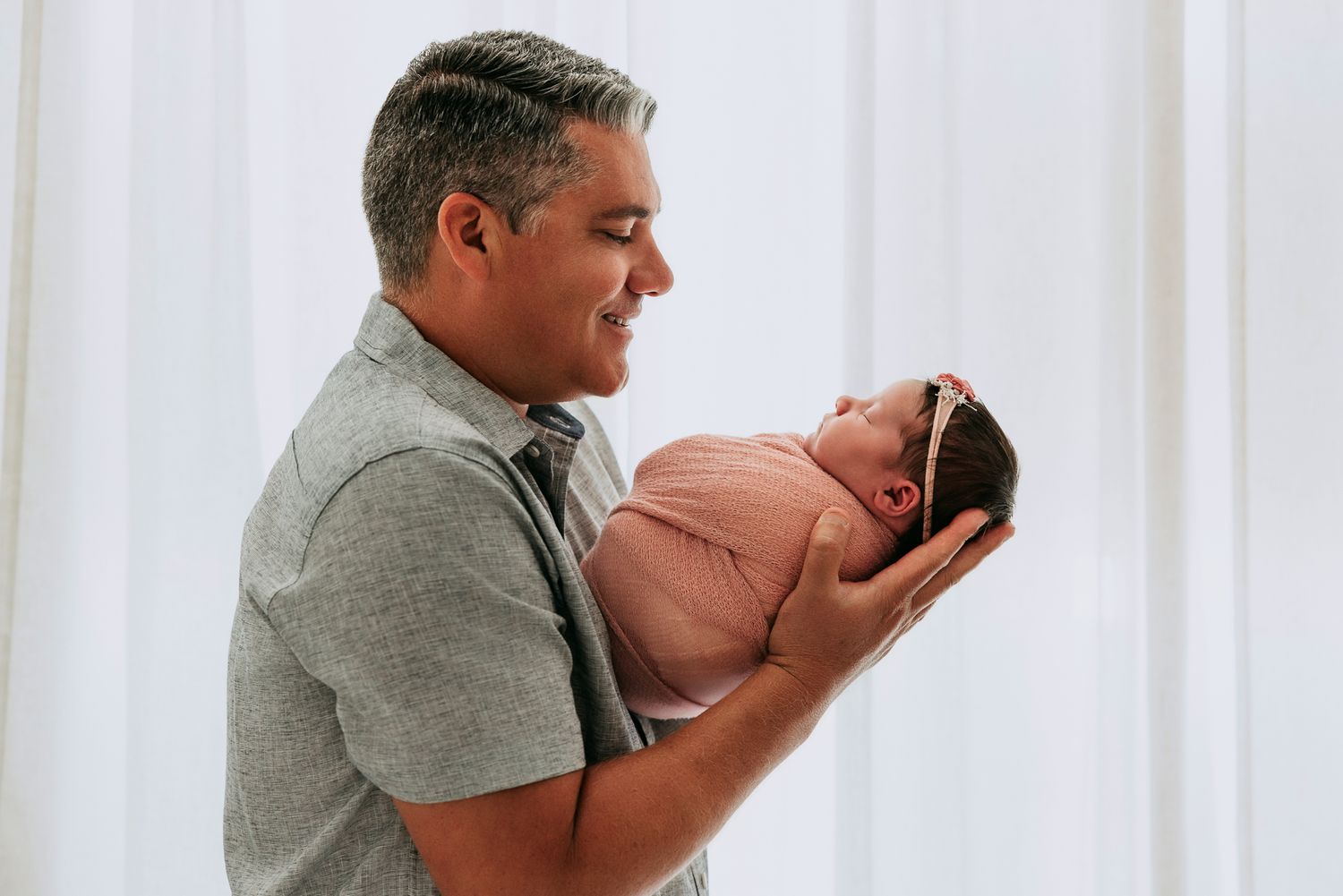 A proud father smiles lovingly while cradling his newborn baby in both hands against a soft white background.
