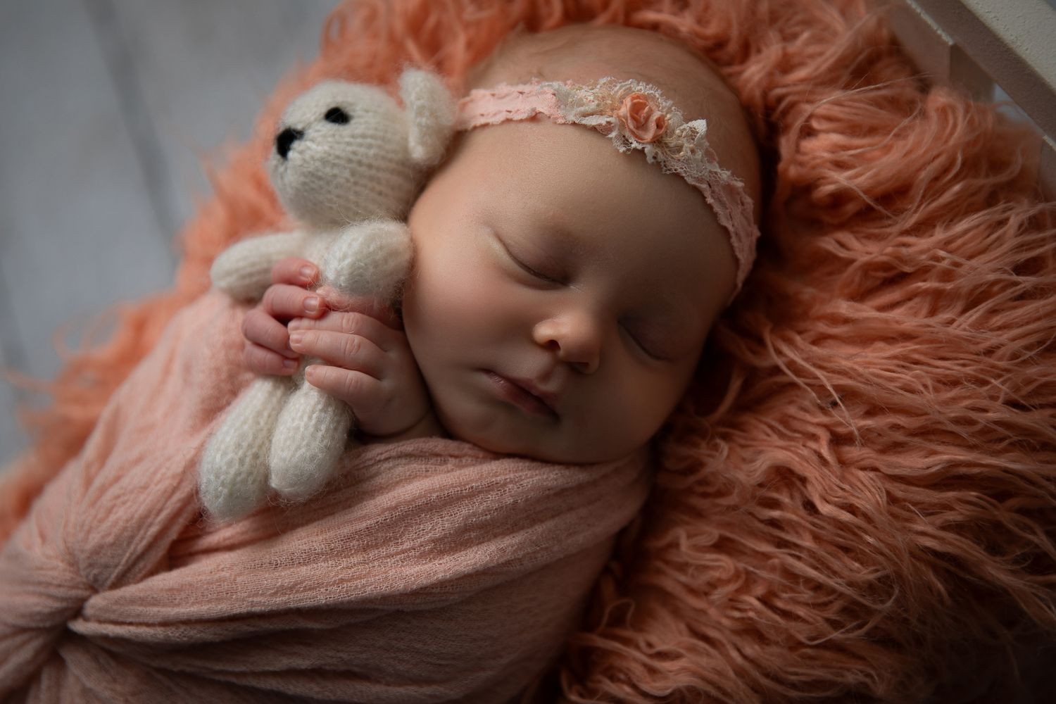 Newborn baby sleeping peacefully wrapped in peach fabric, holding a knit teddy bear on a fluffy orange blanket.