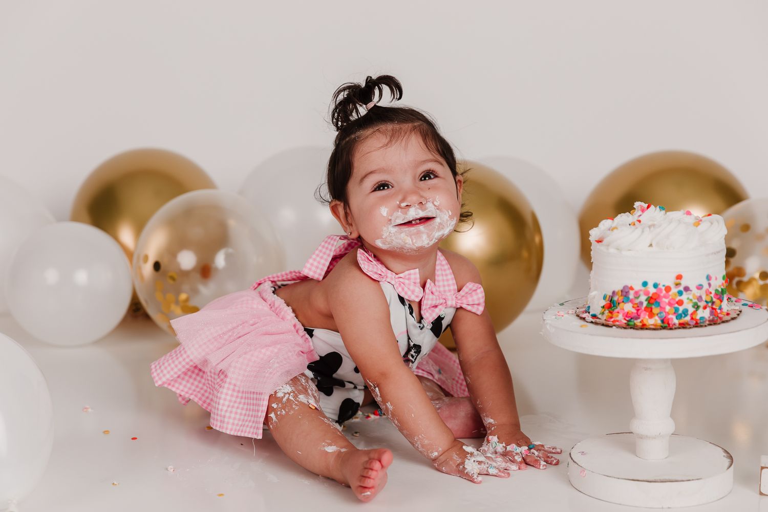 Happy toddler girl in pink dress smashing first birthday cake with balloons.