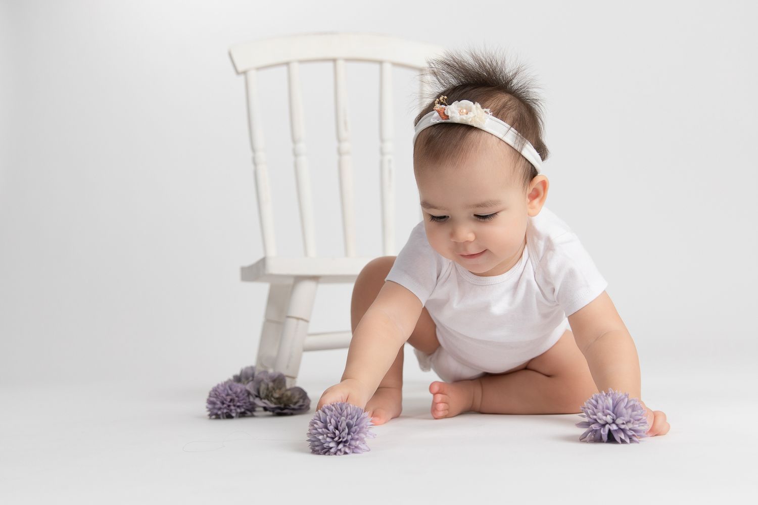 Baby girl in white onesie with headband reaching for purple pom pom toys.