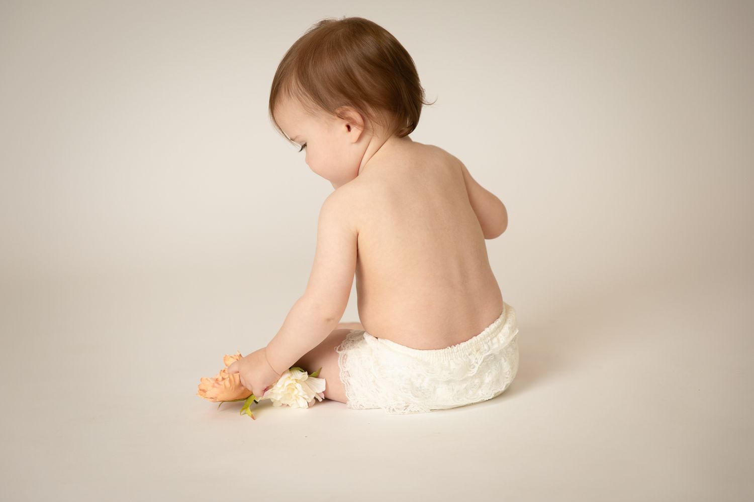 Baby sitting in diaper playing with a flower, photographed from behind on white background.